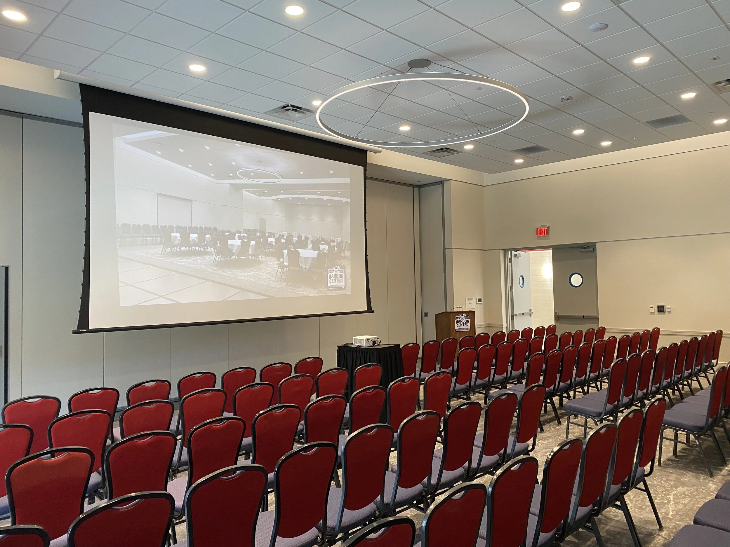 Empty conference room with red chairs facing a large projection screen displaying a ballroom with chairs and tables, with a small podium to the side.