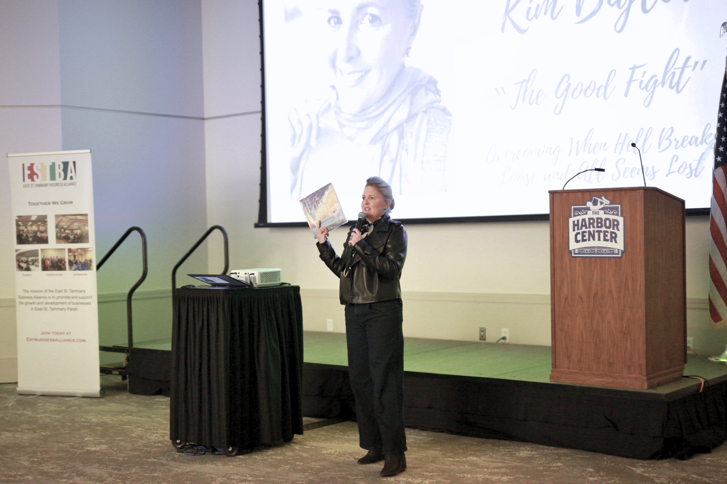 A woman in black clothing is speaking into a microphone and holding a book during a presentation at a conference. There is a large projection screen behind her displaying a woman's face and text, and a podium labelled 'Harbor Center' beside her. To h