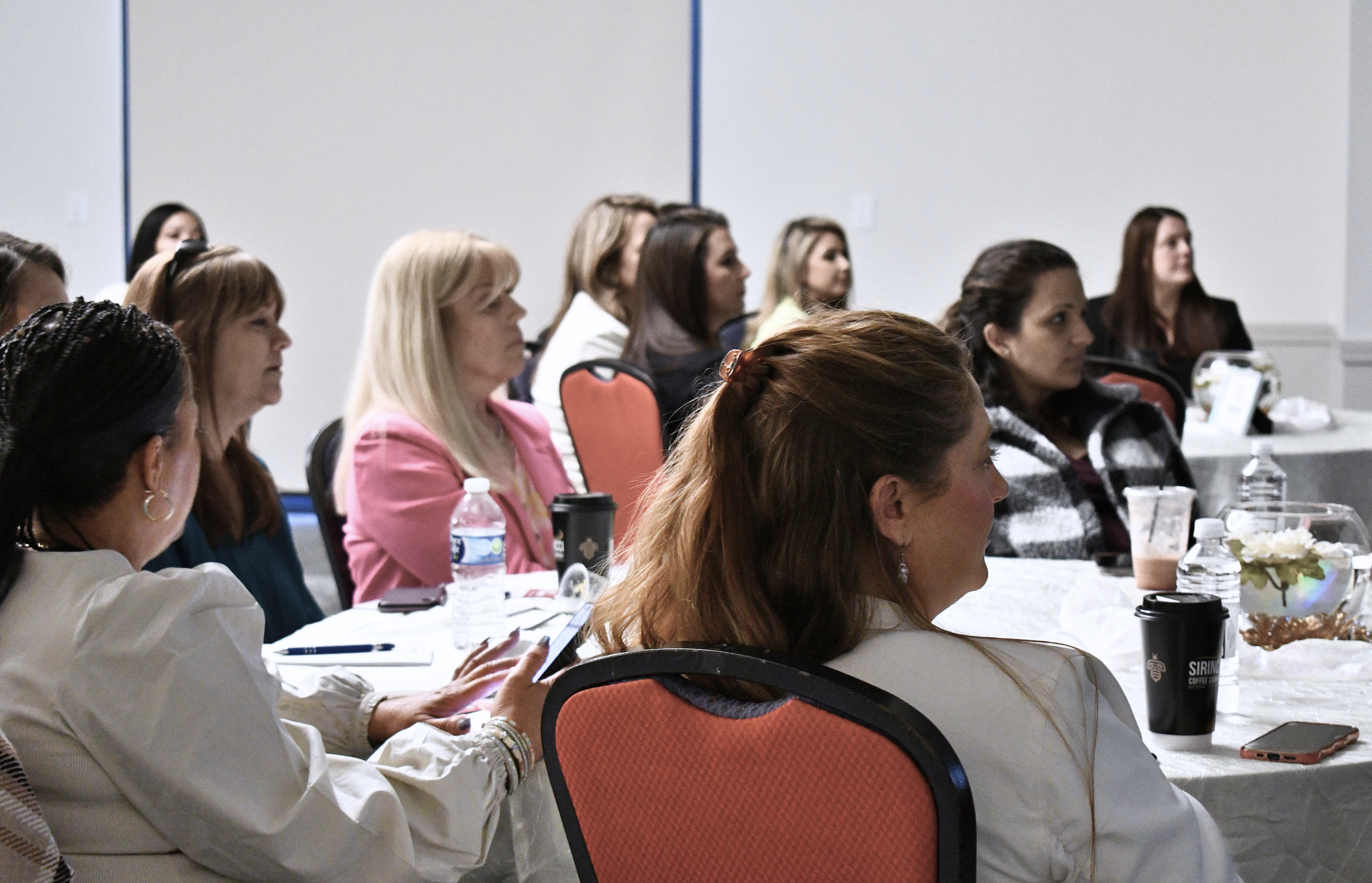 A group of women sitting at round tables during a conference or seminar, listening attentively. The tables have water bottles, coffee cups, and decorative flowers.