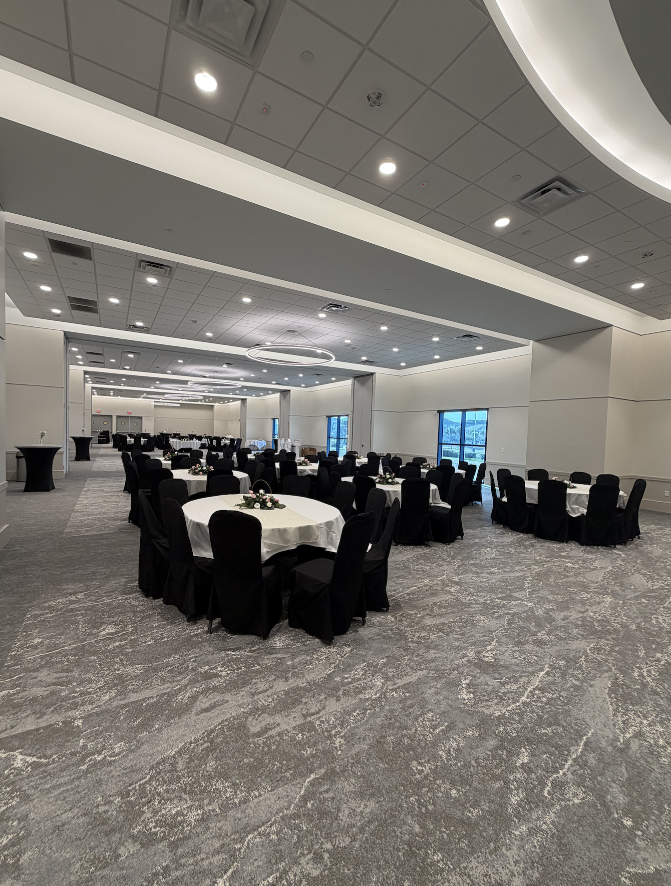 A large banquet hall decorated for an event with round tables covered in white tablecloths and black chairs, with small floral centerpieces on each table. The room has high ceilings with circular modern light fixtures, large windows on the side, and 