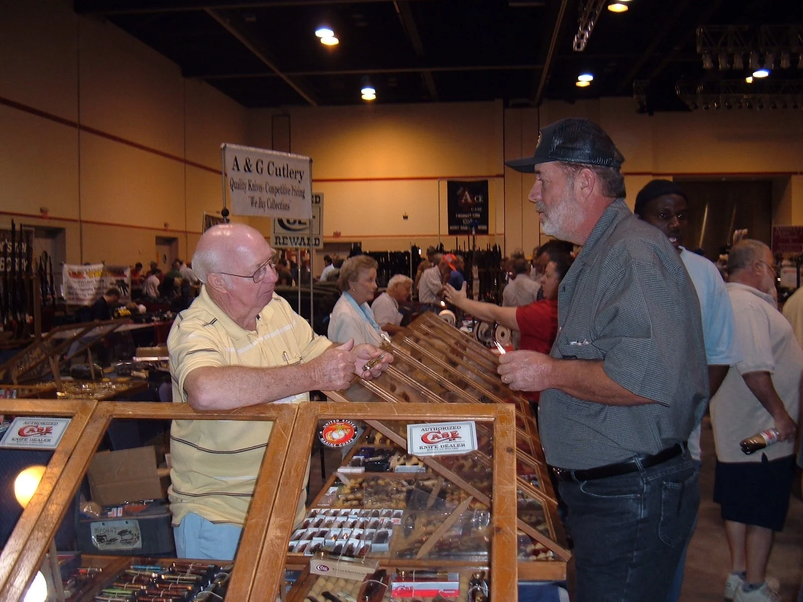 A man with a gray beard and wearing a cap and gray shirt shopping at a knife booth. He is talking to an older man in a yellow striped shirt behind the display case, which contains various knives and cutlery. The background shows a busy indoor event w