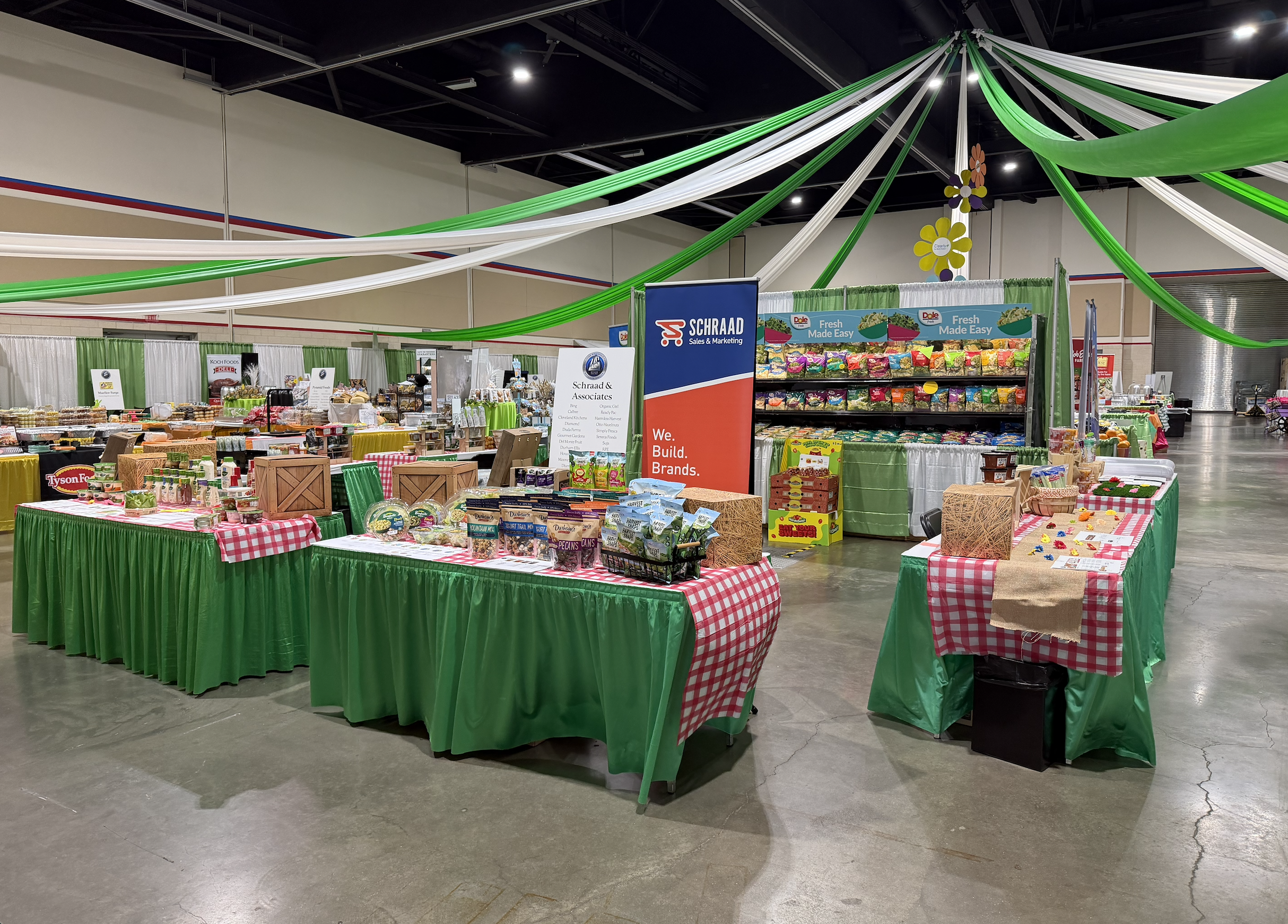Indoor market or trade show with tables covered in green and red checkered tablecloths displaying various products, a promotional banner for Schraad Sales & Marketing, shelves stocked with snacks, and decorative green and white streamers hanging from