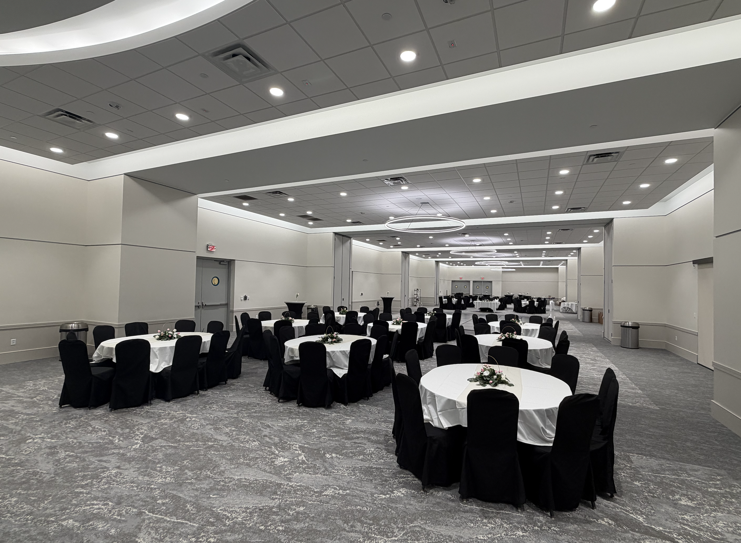 An empty banquet hall with round tables covered with white tablecloths and black chairs. Each table has a floral centerpiece and a beige table runner. The ceiling has recessed lighting and modern circular chandeliers. The room has a gray patterned ca