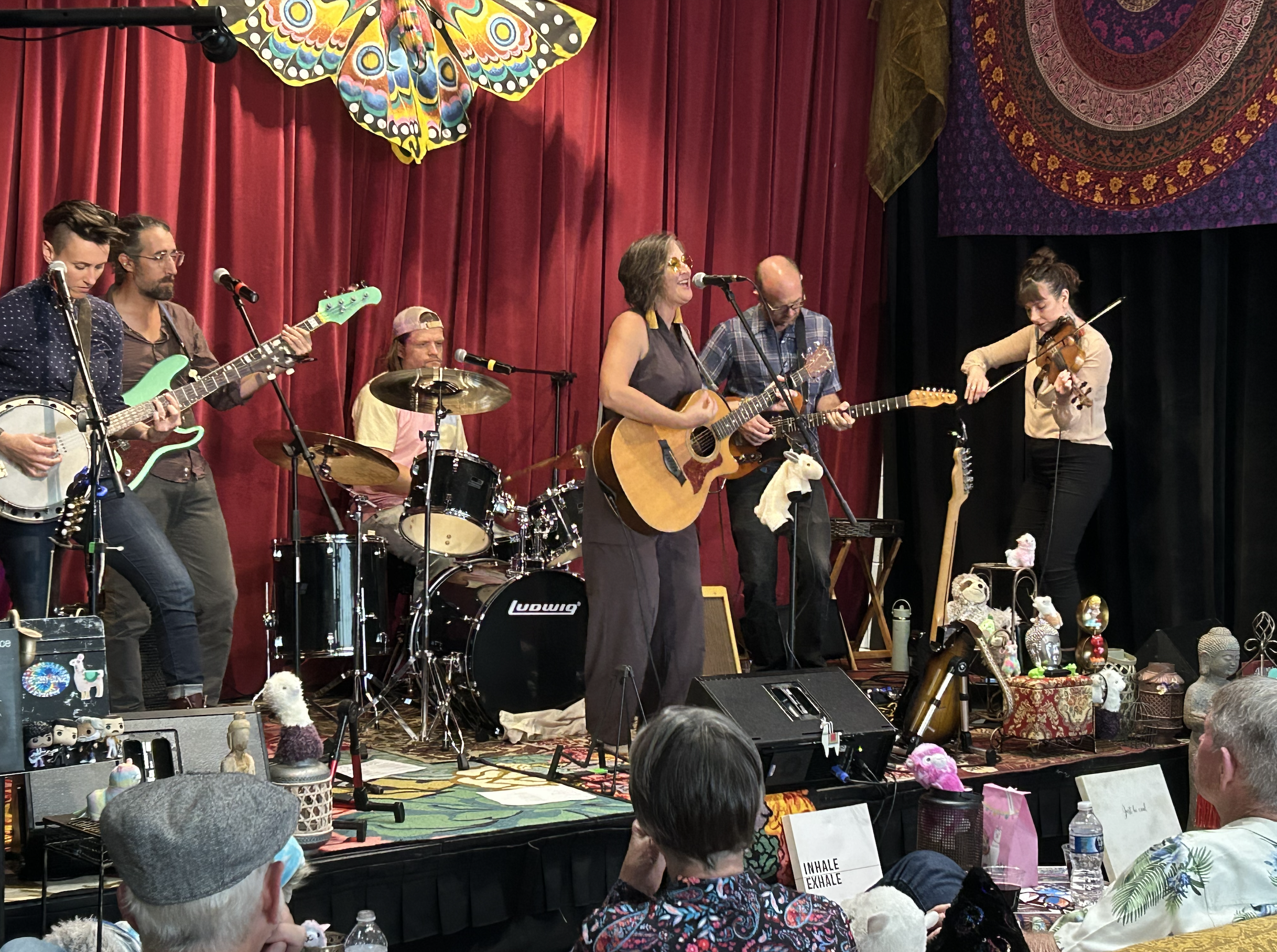 A band performing live on stage with a red curtain backdrop, featuring six musicians playing various instruments including guitars, drums, and a violin.