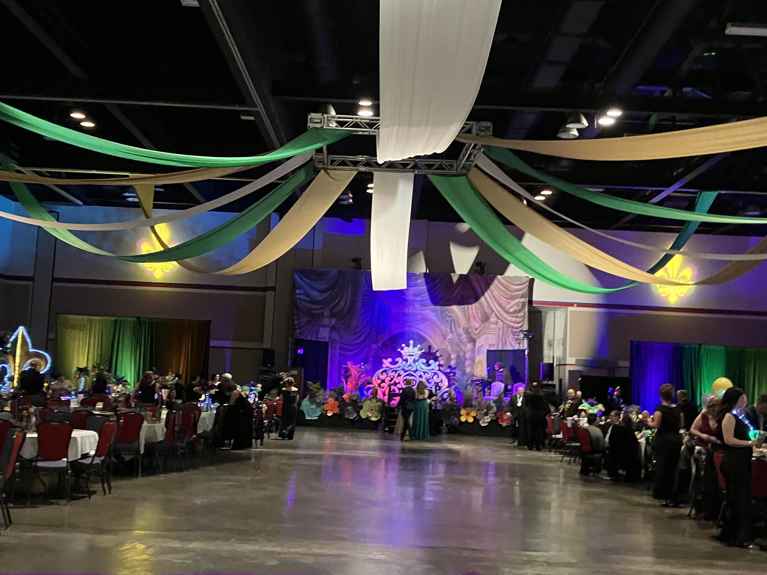 A decorated banquet hall with tables and chairs, people dressed formally, and a stage with colorful lighting and floral decorations at an event or celebration.