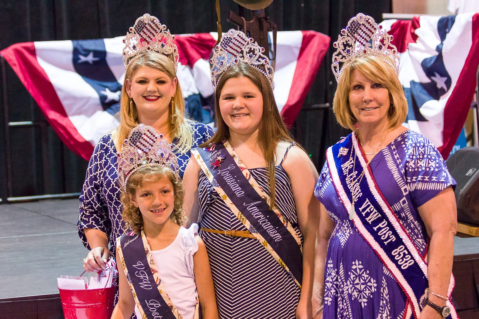 Group of five women and girls wearing tiaras and sashes celebrating, with an American flag-themed backdrop behind them.