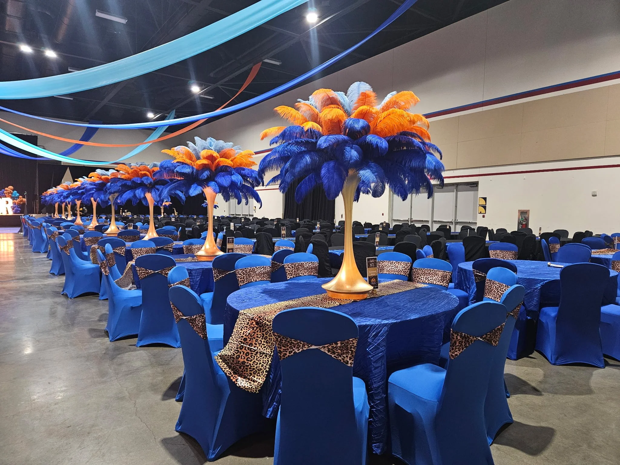 Event hall decorated with blue and leopard print accents, featuring long tables with blue tablecloths, blue chairs with leopard print chair bands, and large feather trees in blue, orange, and gold.