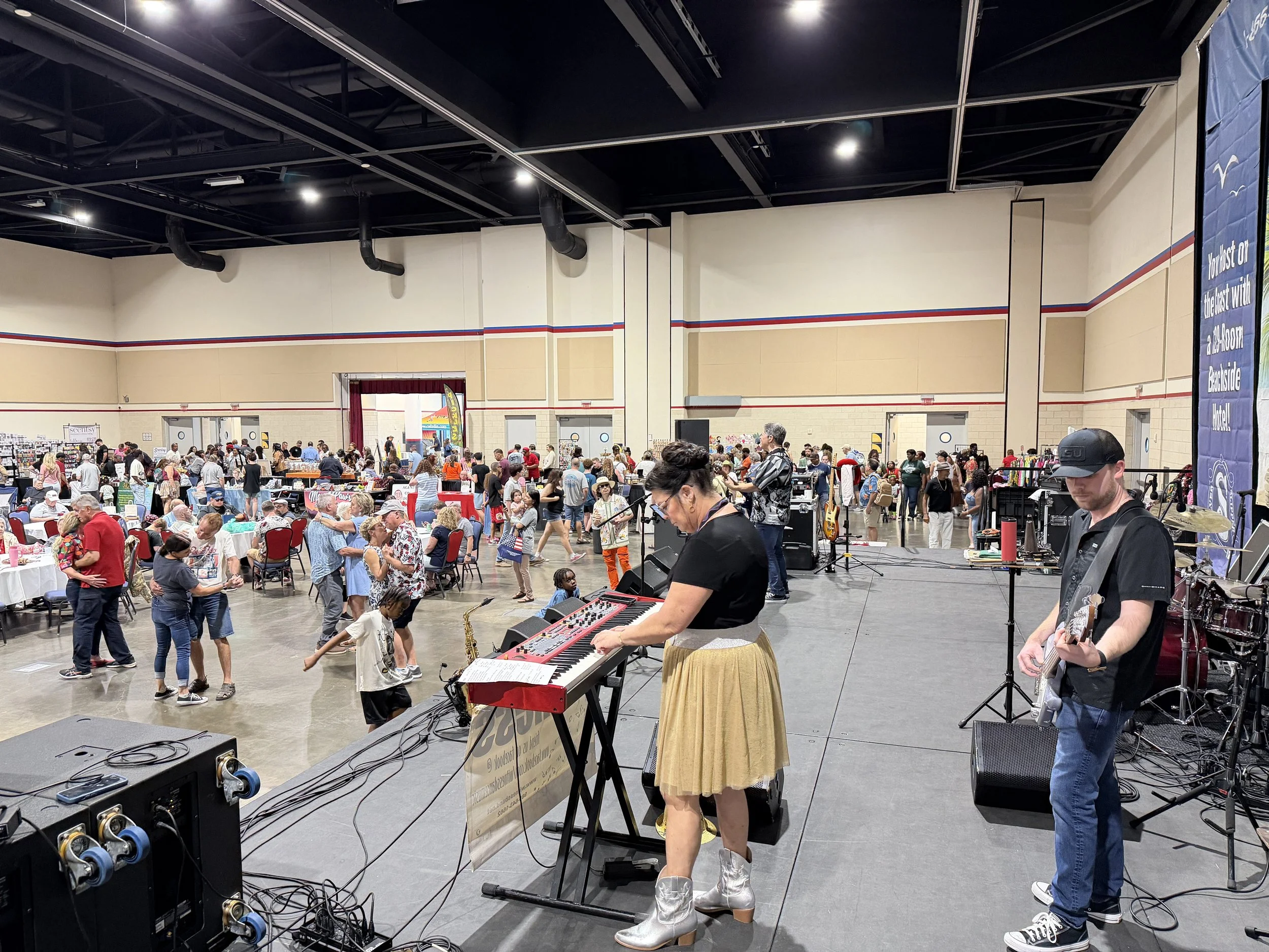Live music performance with a female keyboardist and male guitarist on a stage in a crowded indoor event with people dancing and socializing, and tables with seated guests in the background.