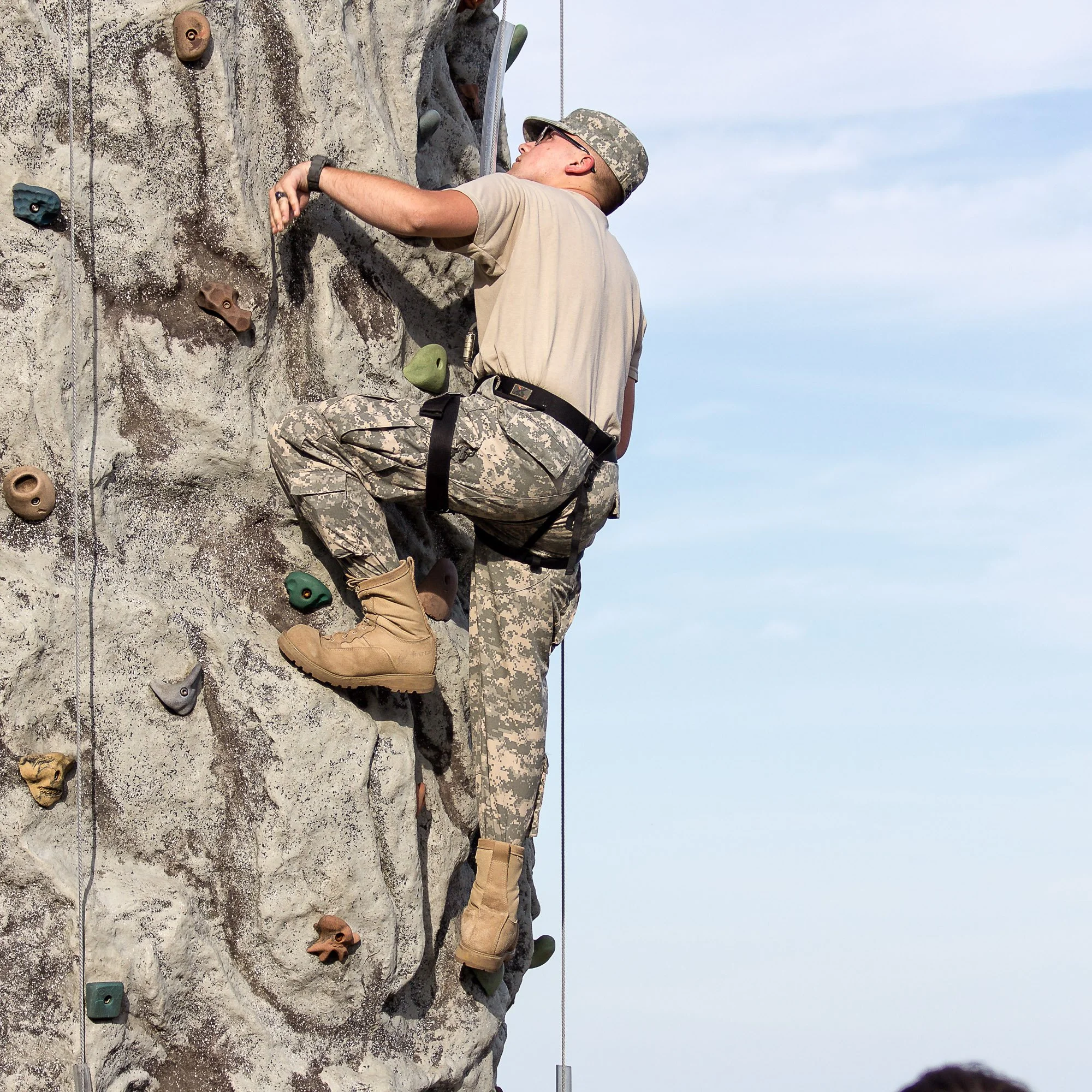 A soldier climbing an outdoor rock wall with colorful handholds and footholds during daytime.