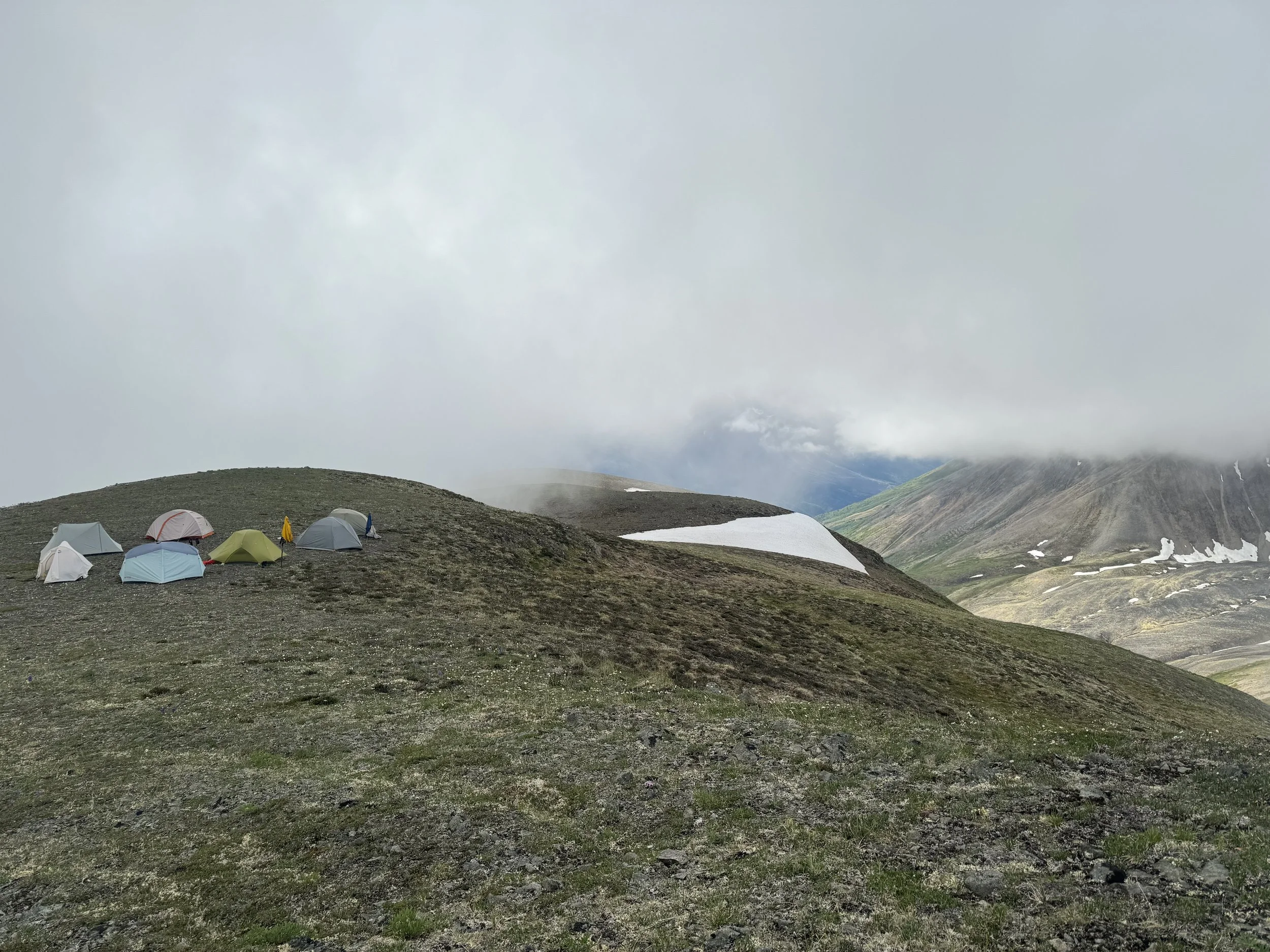 Several tents pitched on a mountain slope with snow patches and cloudy sky in the background.
