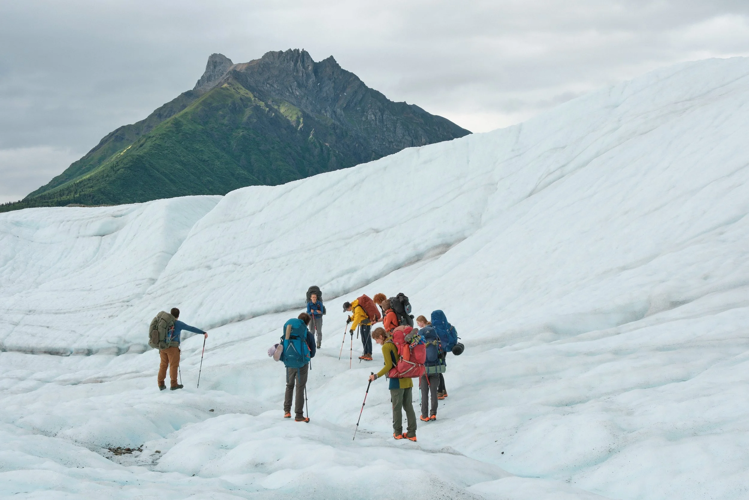 A serene landscape featuring a turquoise lake with a rocky shore, green and brown hills, and snow-capped mountains in the background under a partly cloudy blue sky.