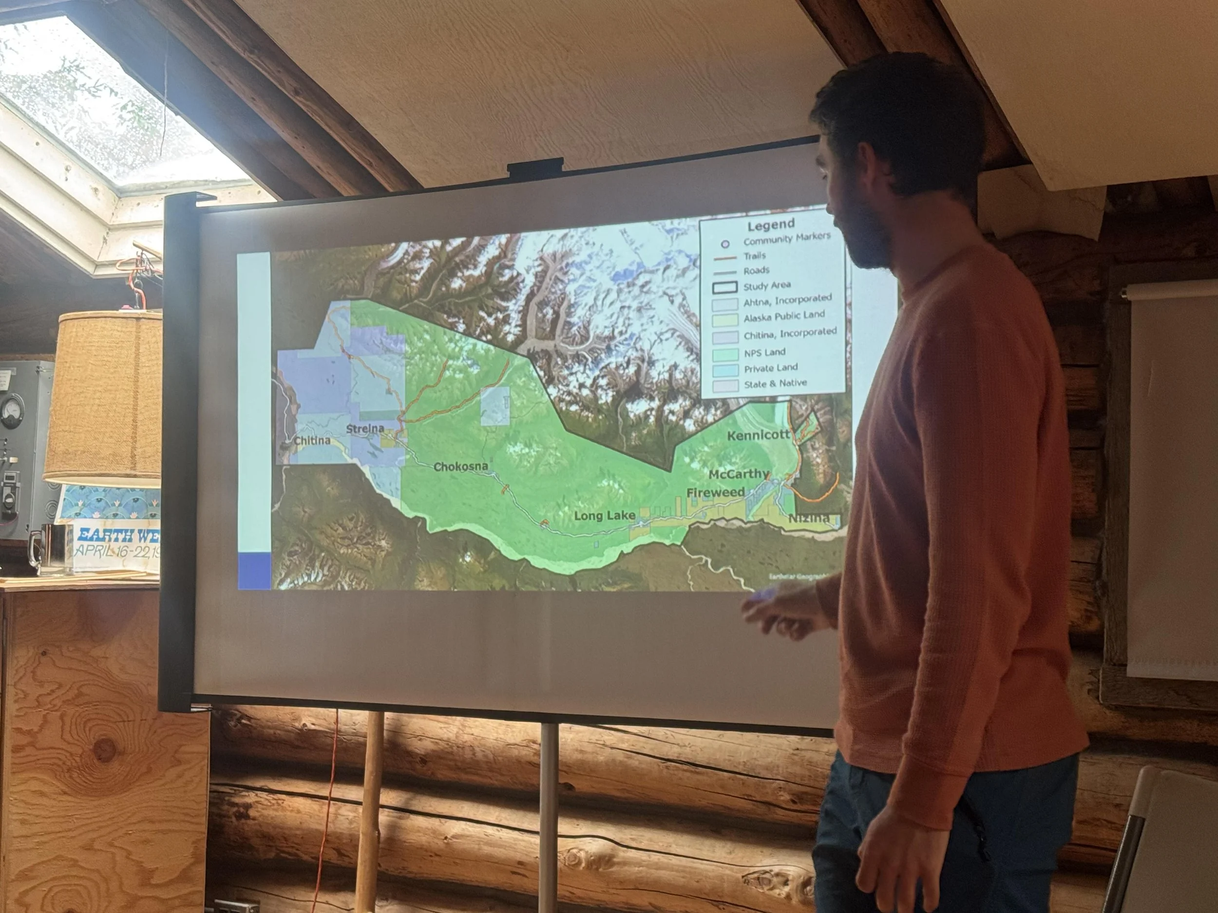 Man in salmon sweater giving presentation, map on screen showing land and water, legend with land types, community markers, trails, roads, and study areas.