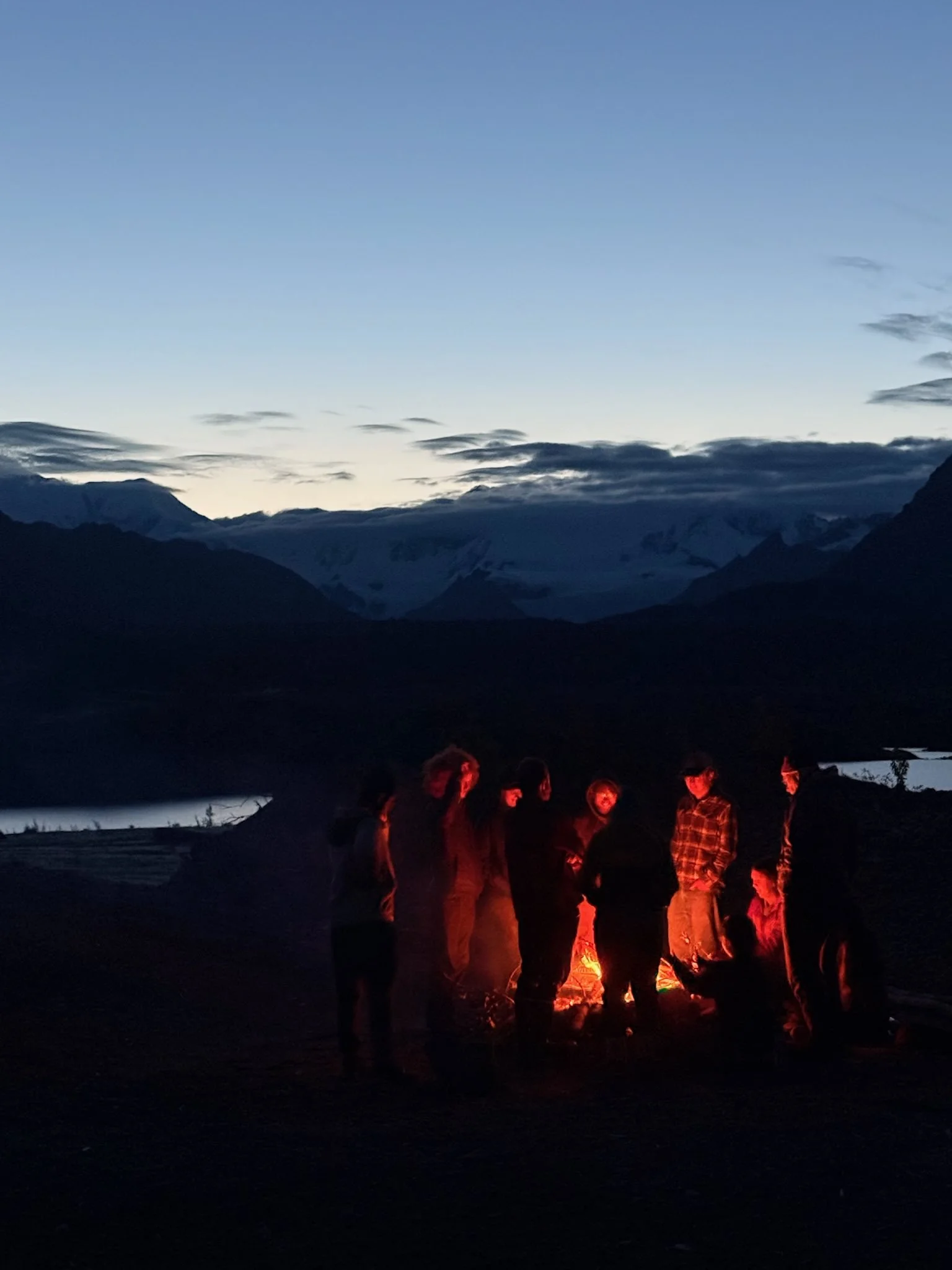 Group of people gathered around a campfire outdoors during dusk, with mountains and a lake in the background.