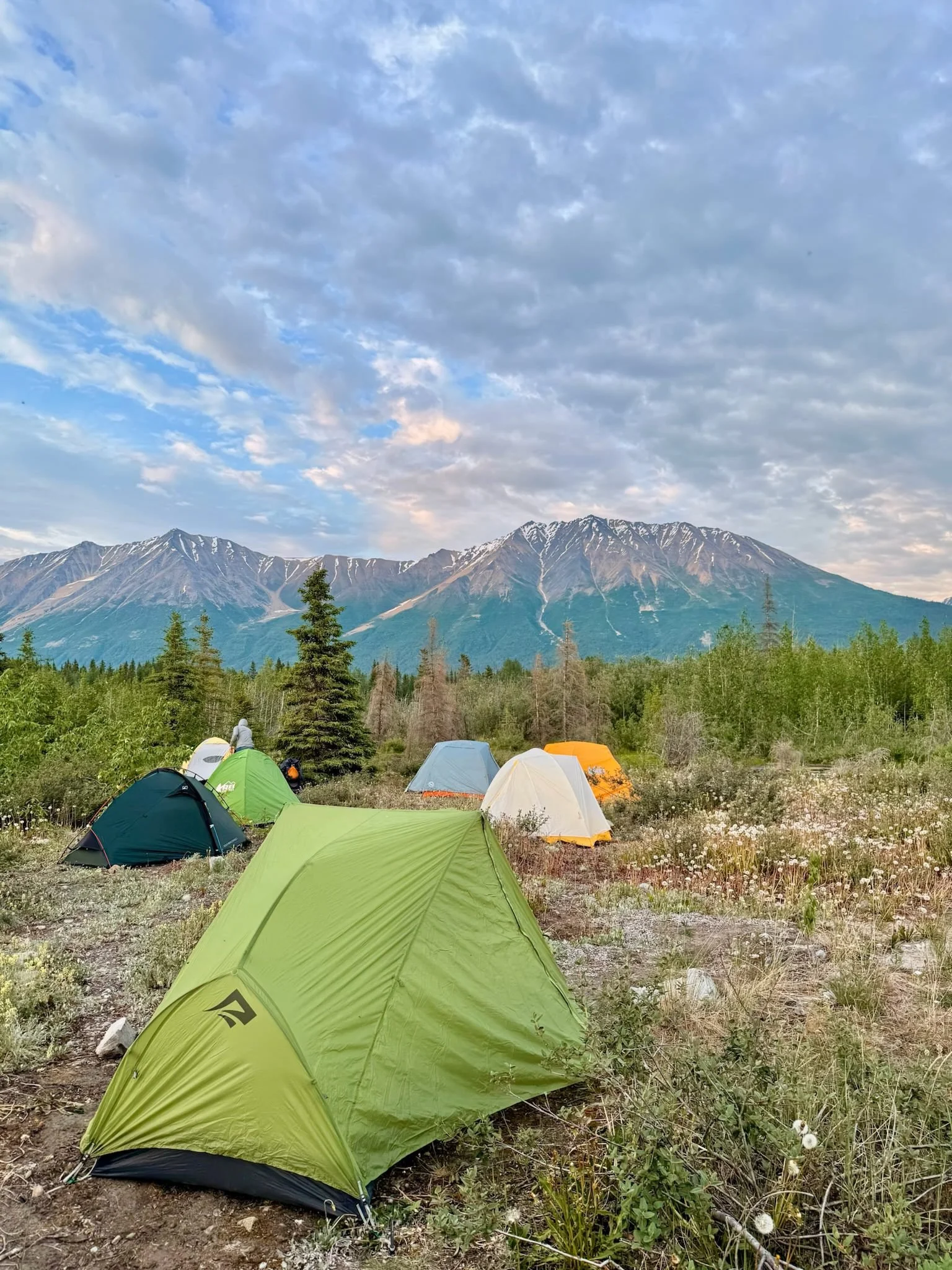 A campsite with several colored tents set up in a grassy clearing, with mountains in the background and a sky filled with clouds.
