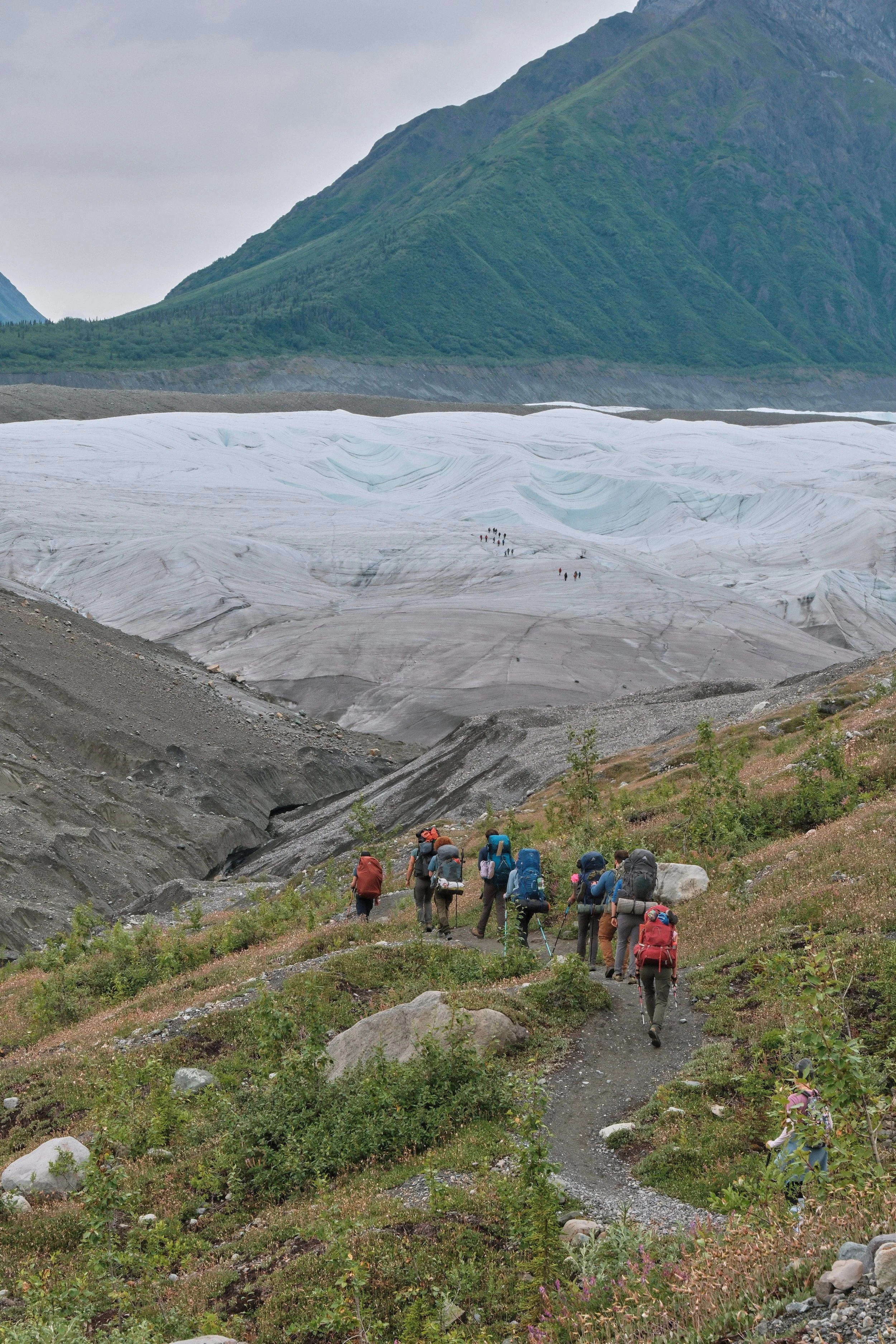 A group of hikers walking along a trail in a mountainous area with a glacier in the background.