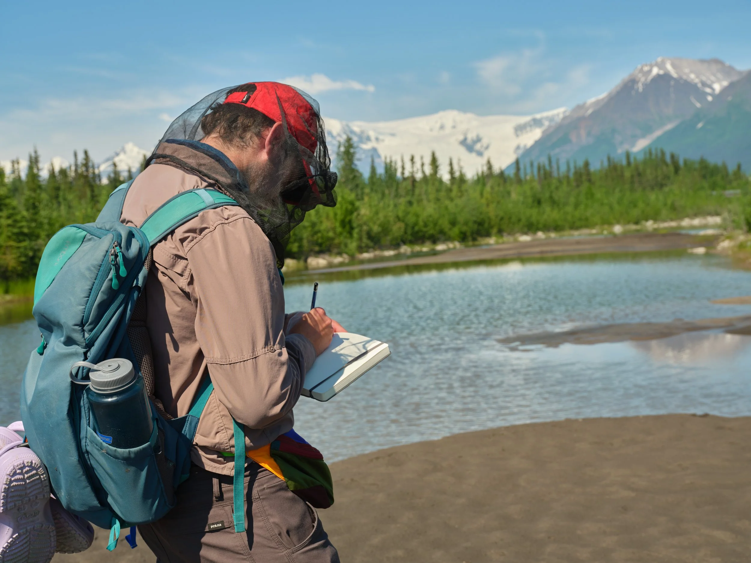 Two people sitting on rocky ground in front of a glacier and snow-capped mountains, one pointing at the glacier.