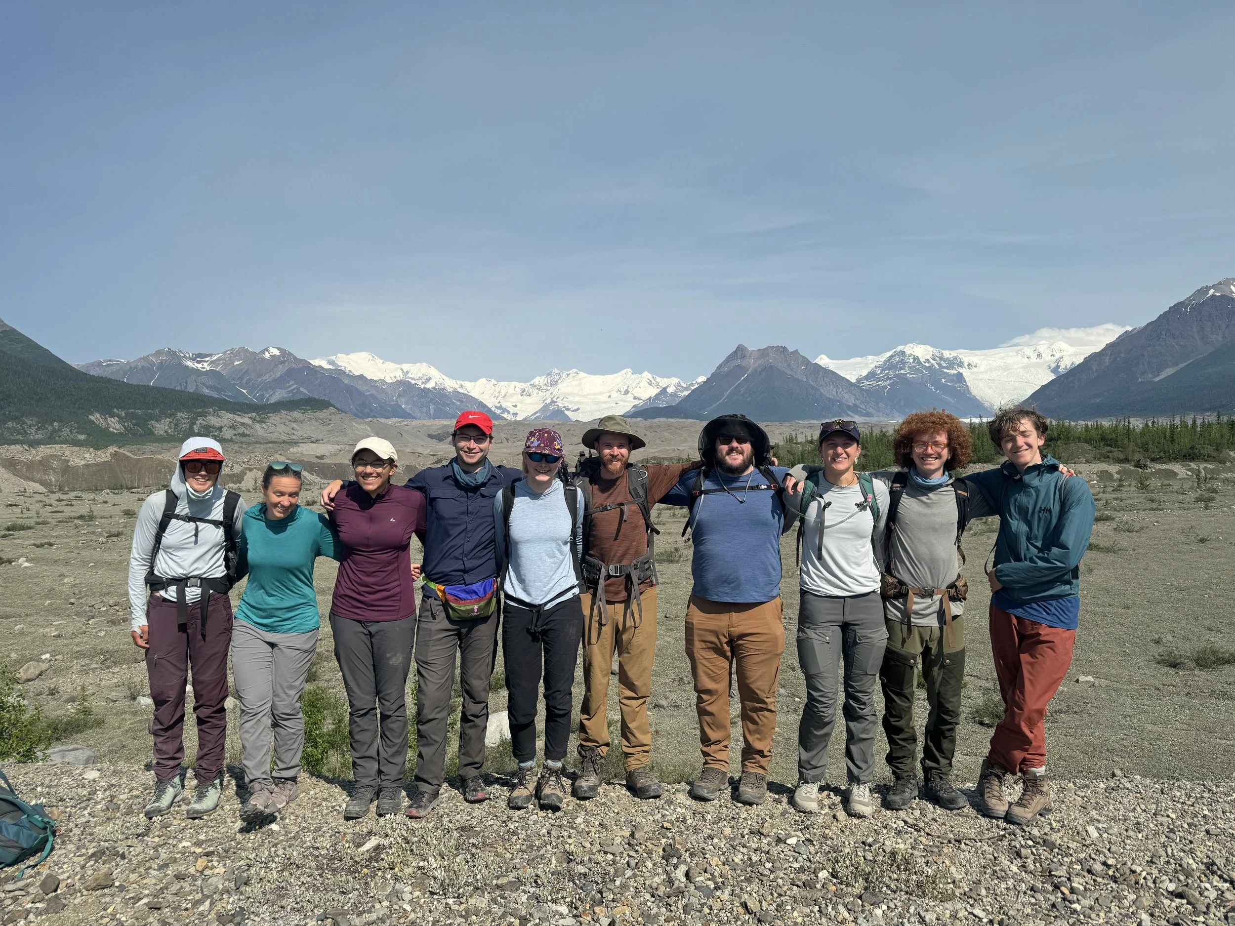 Group of nine hikers standing outdoors in front of mountains with snow-capped peaks, wearing backpacks and outdoor clothing.