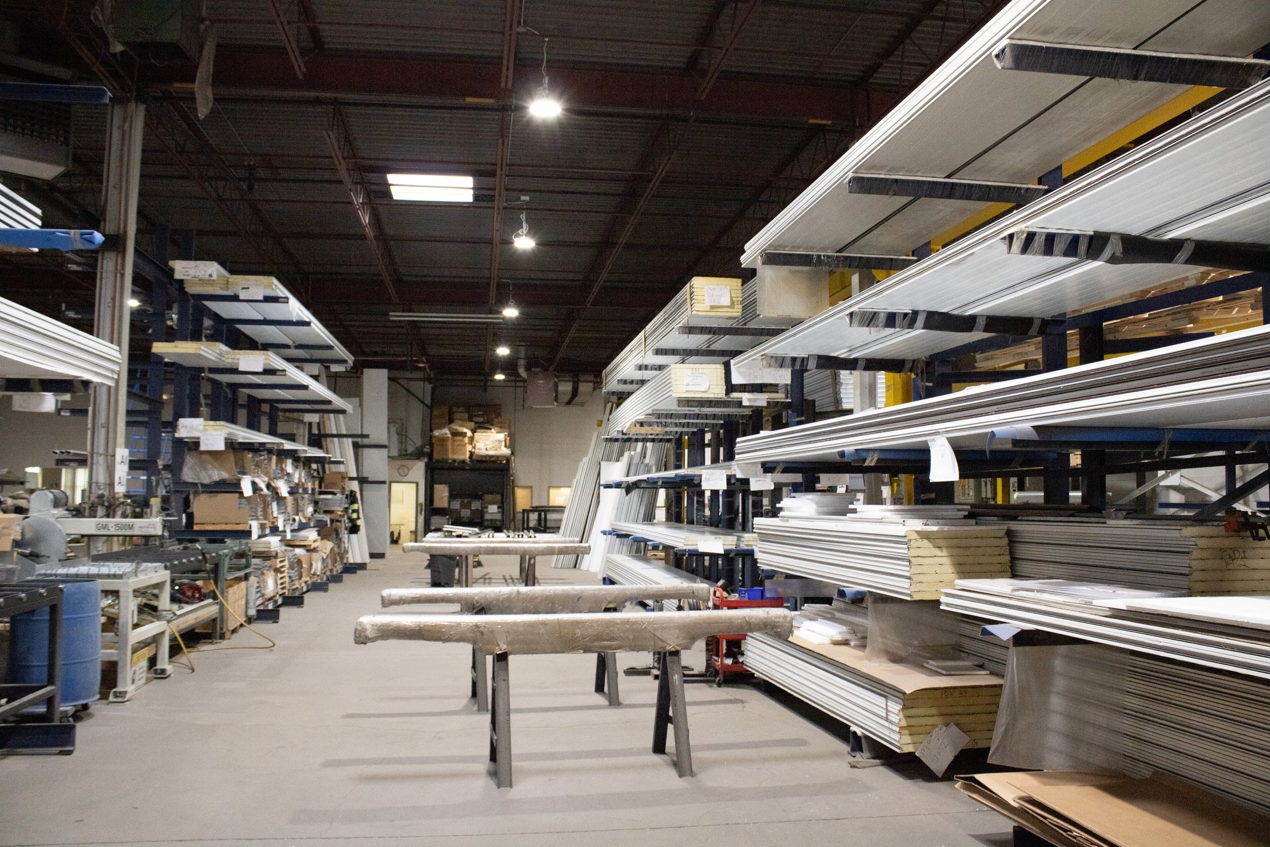 Interior of a hardware store or warehouse with metal shelves stocked with wood, metal, and other building materials, and workbenches in the foreground.