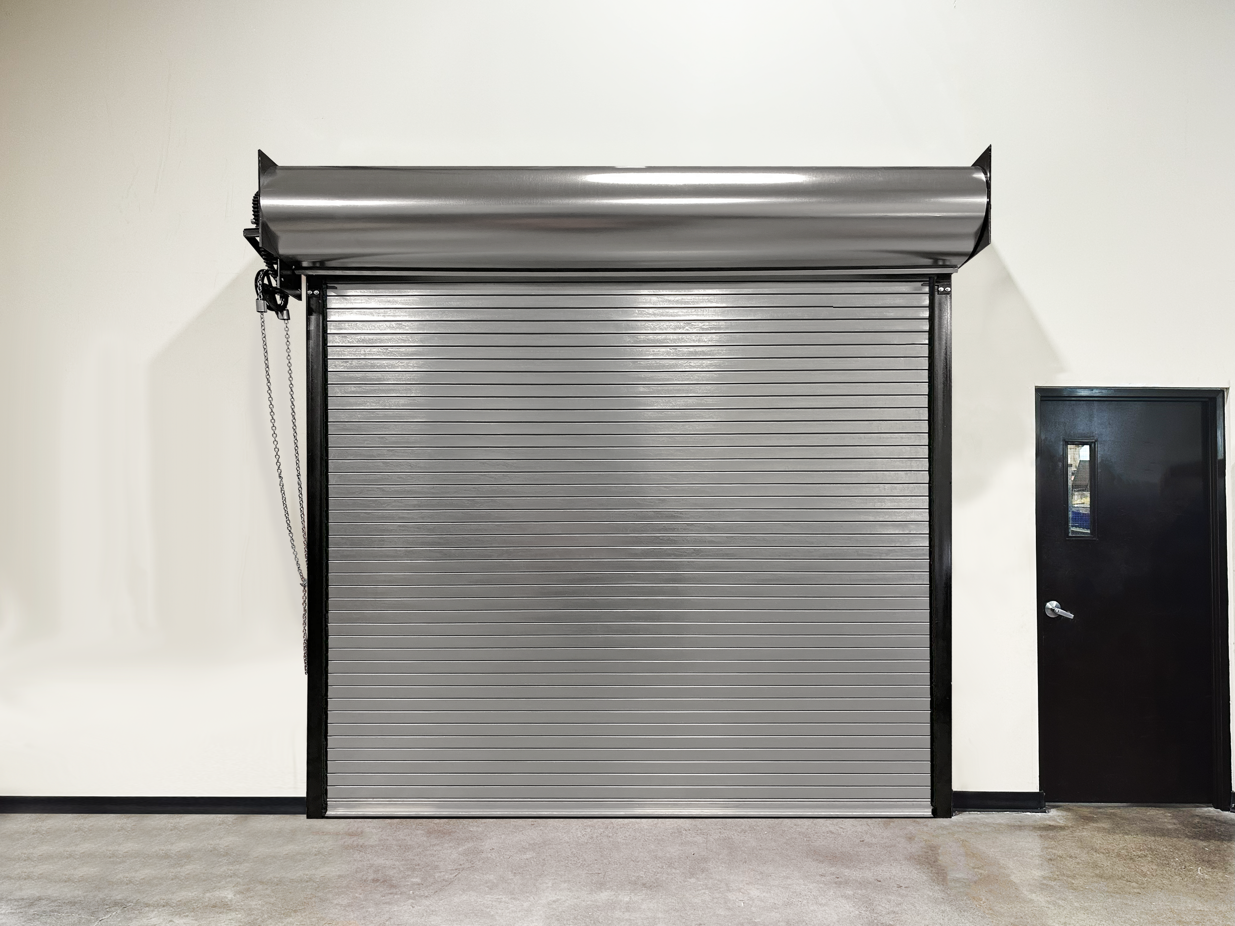 A metallic gray rolling shutter door closed against a white wall in a plain room with a dark door on the right.