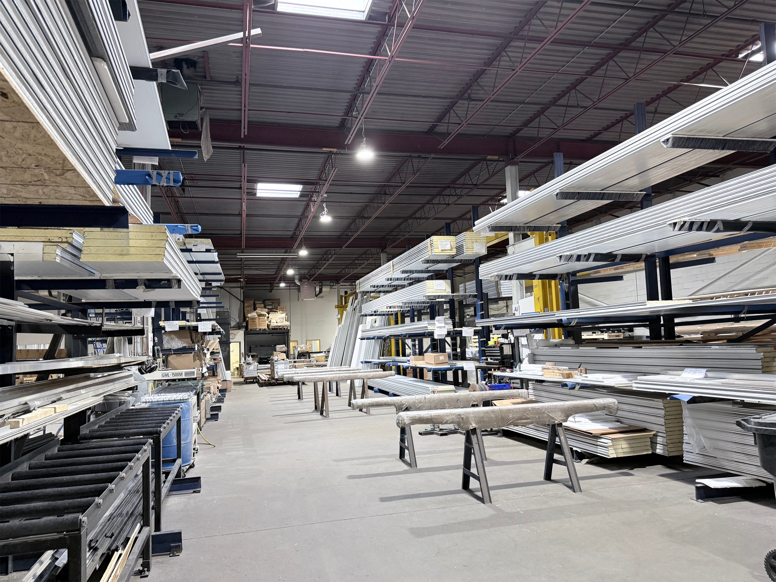 Interior of a warehouse store with metal shelves filled with various building materials such as metal boards, drywall, and pipes. The ceiling is high with visible metal beams and bright overhead lighting.