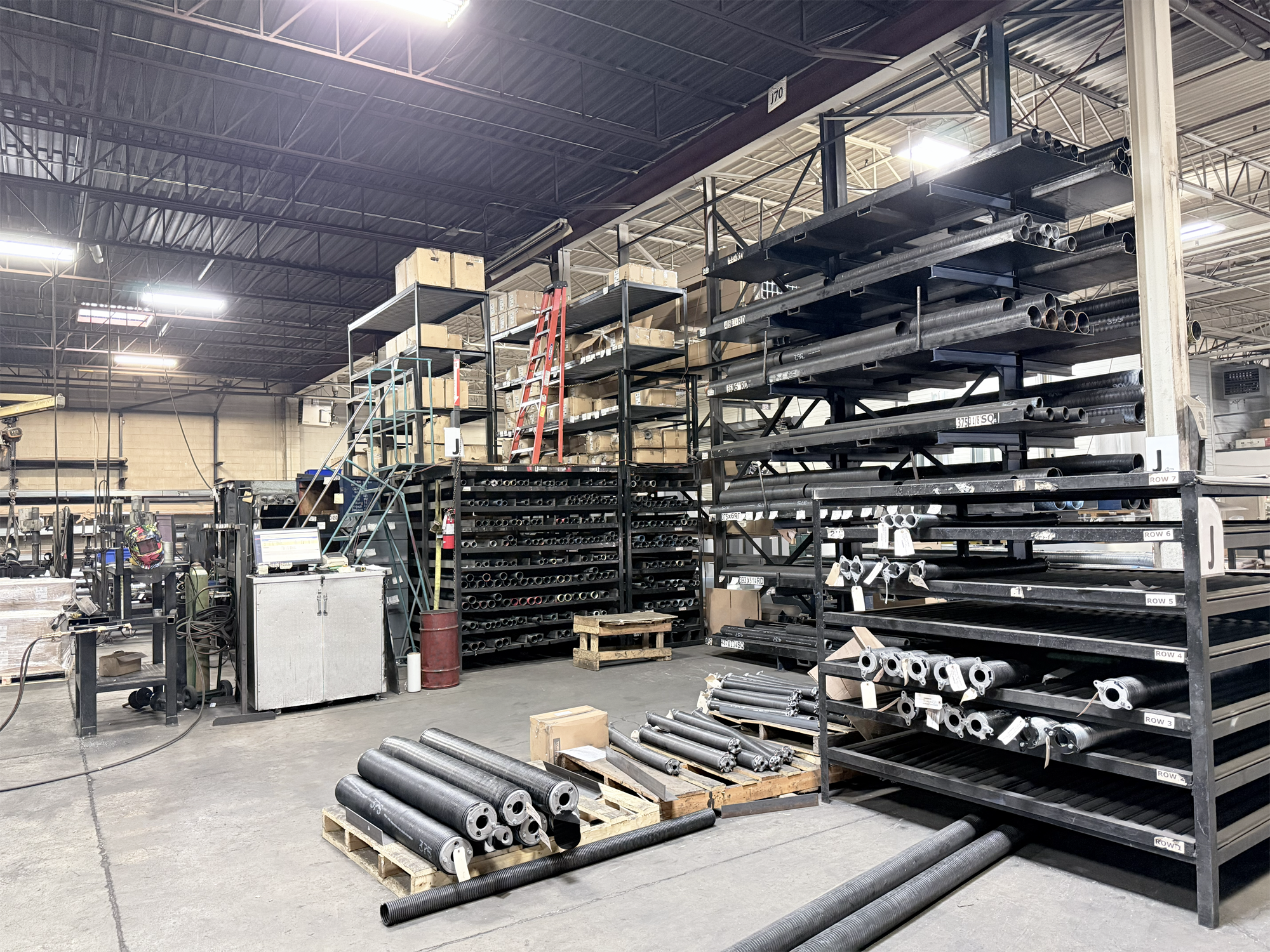Interior of a warehouse industrial storage area with metal pipes and boxes on shelves and pallets, a ladder, and various equipment and supplies scene.