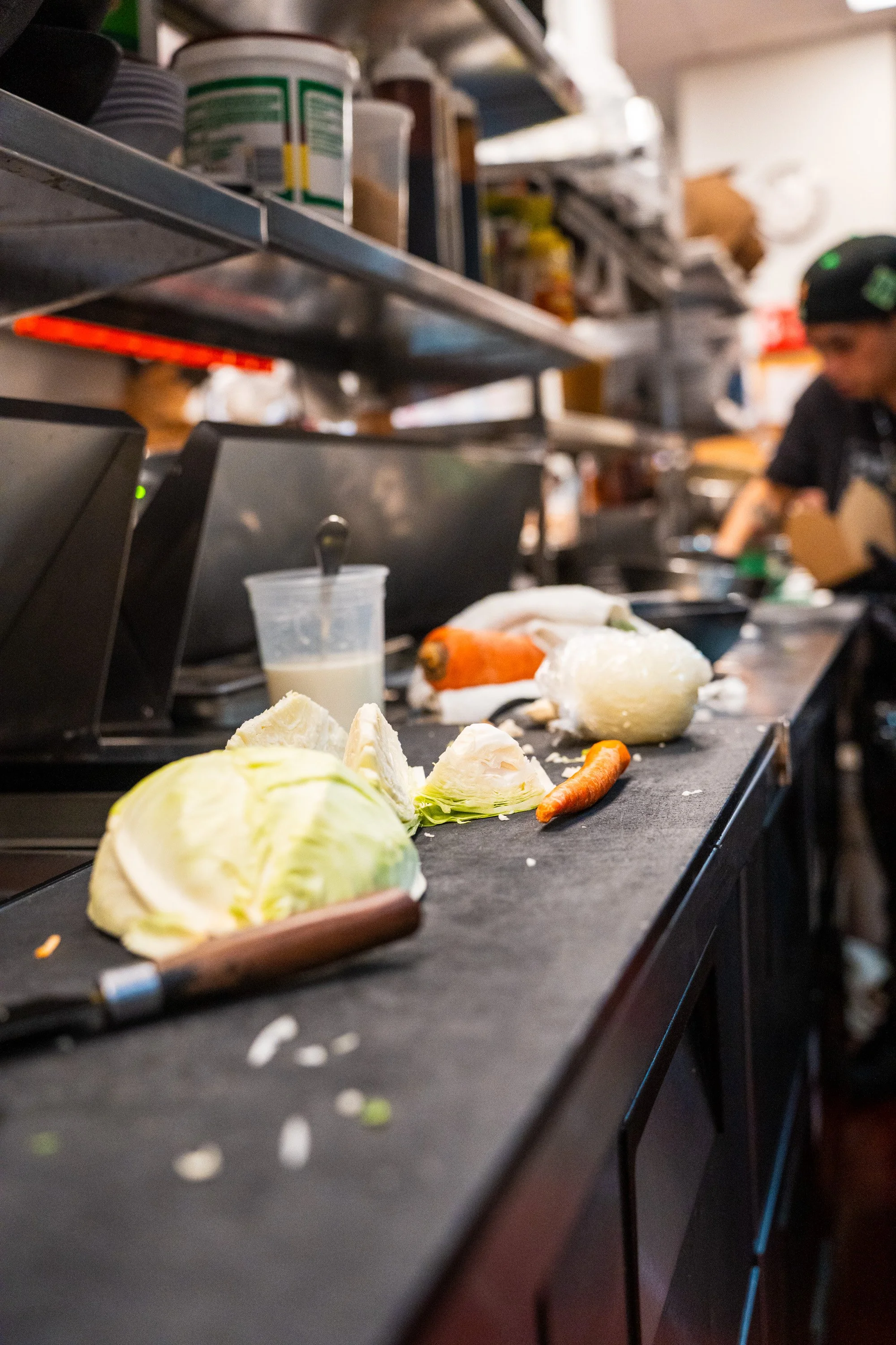 Kitchen countertop with vegetables including cabbage, carrot, and onion, and a clear plastic cup with a white liquid, with a person working in the background.