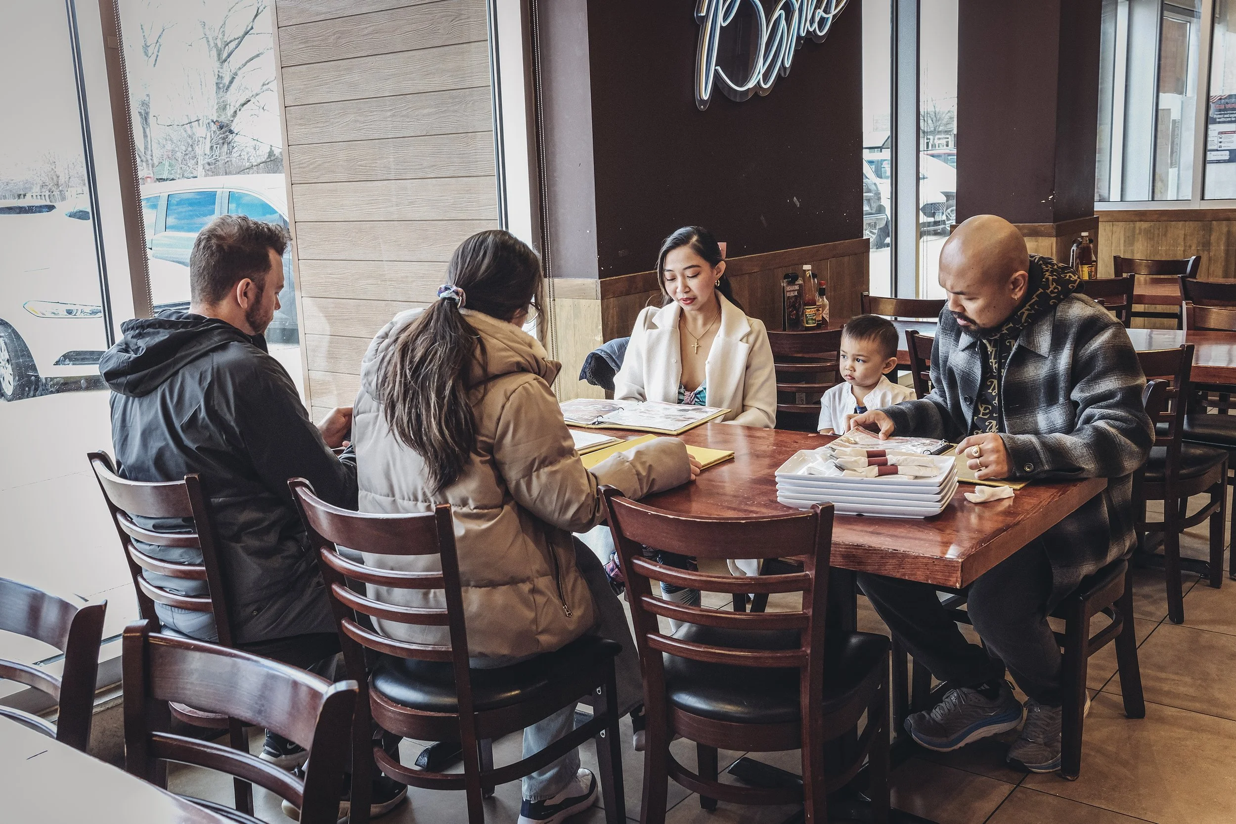 People sitting at a restaurant table near the window, looking at menus, with condiments on the table, and a neon sign on the wall behind them.