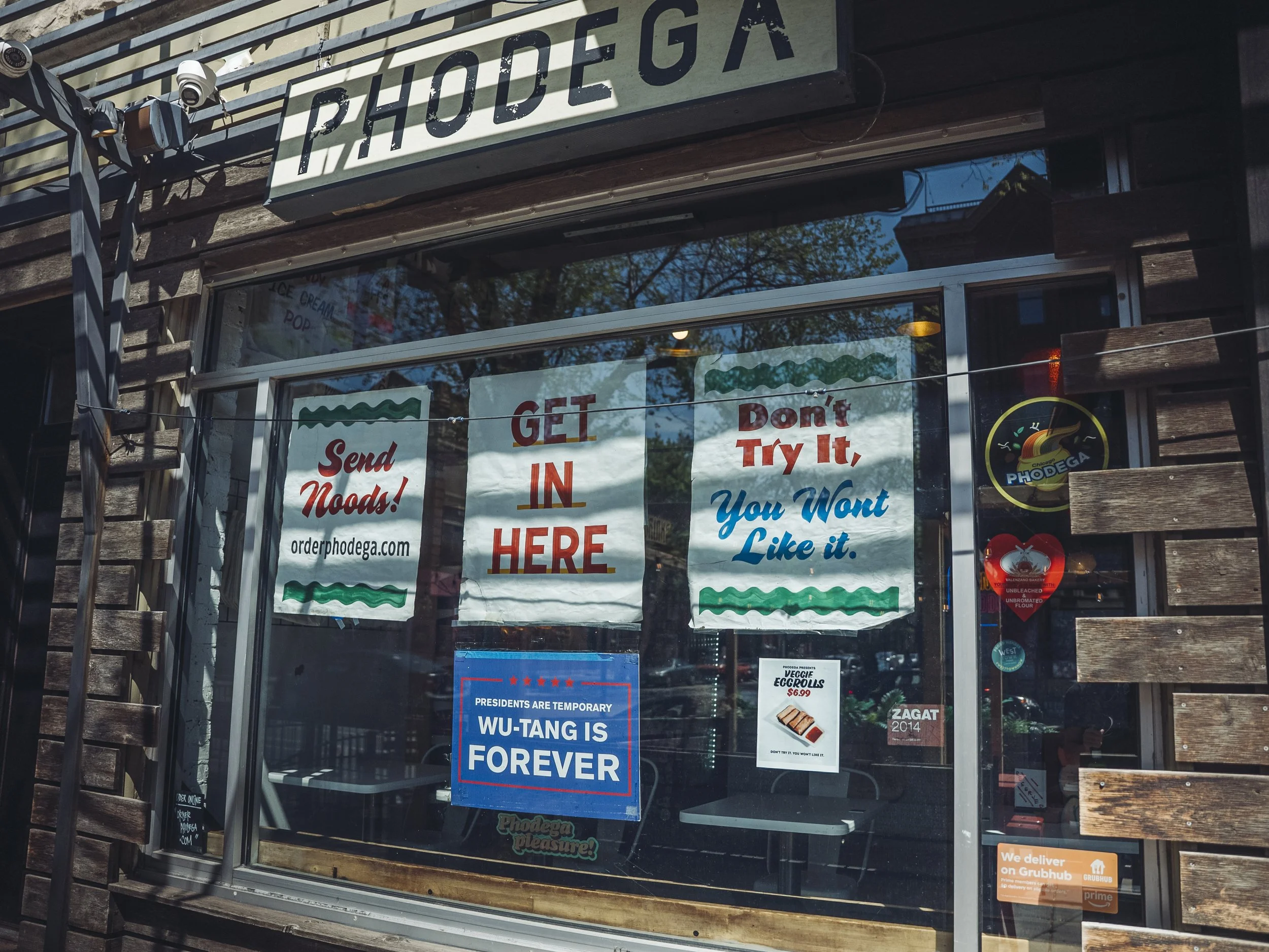 Front window of a restaurant or deli with various signs and posters, including a prominent sign that says 'SEND IN' and other messages about food and promotions, reflected in the glass window with part of the building’s exterior wooden siding visible.