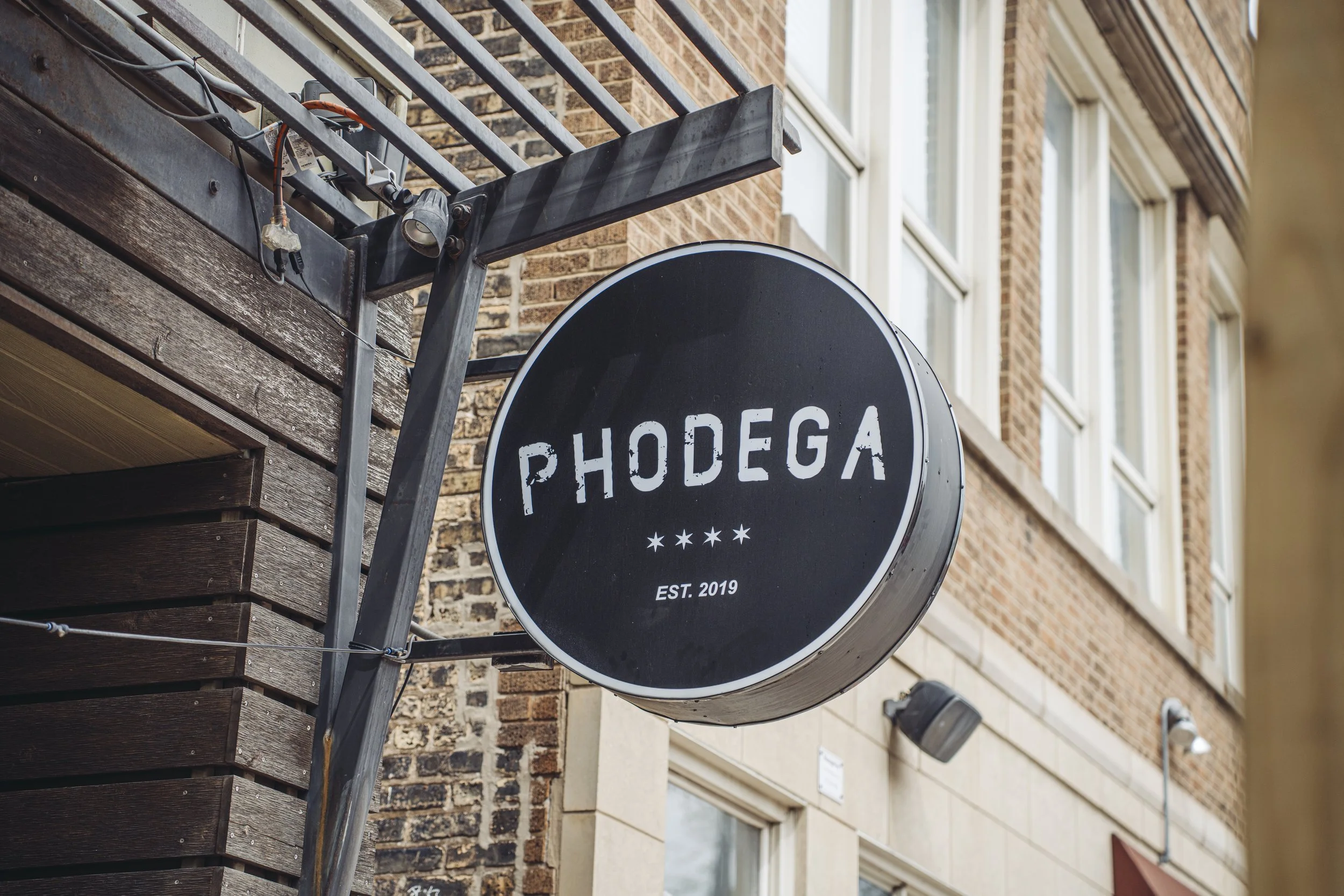A circular black sign with white text reading 'RHODEGA' and 'EST. 2019', attached to the side of a building with brick and wood siding, above a street.