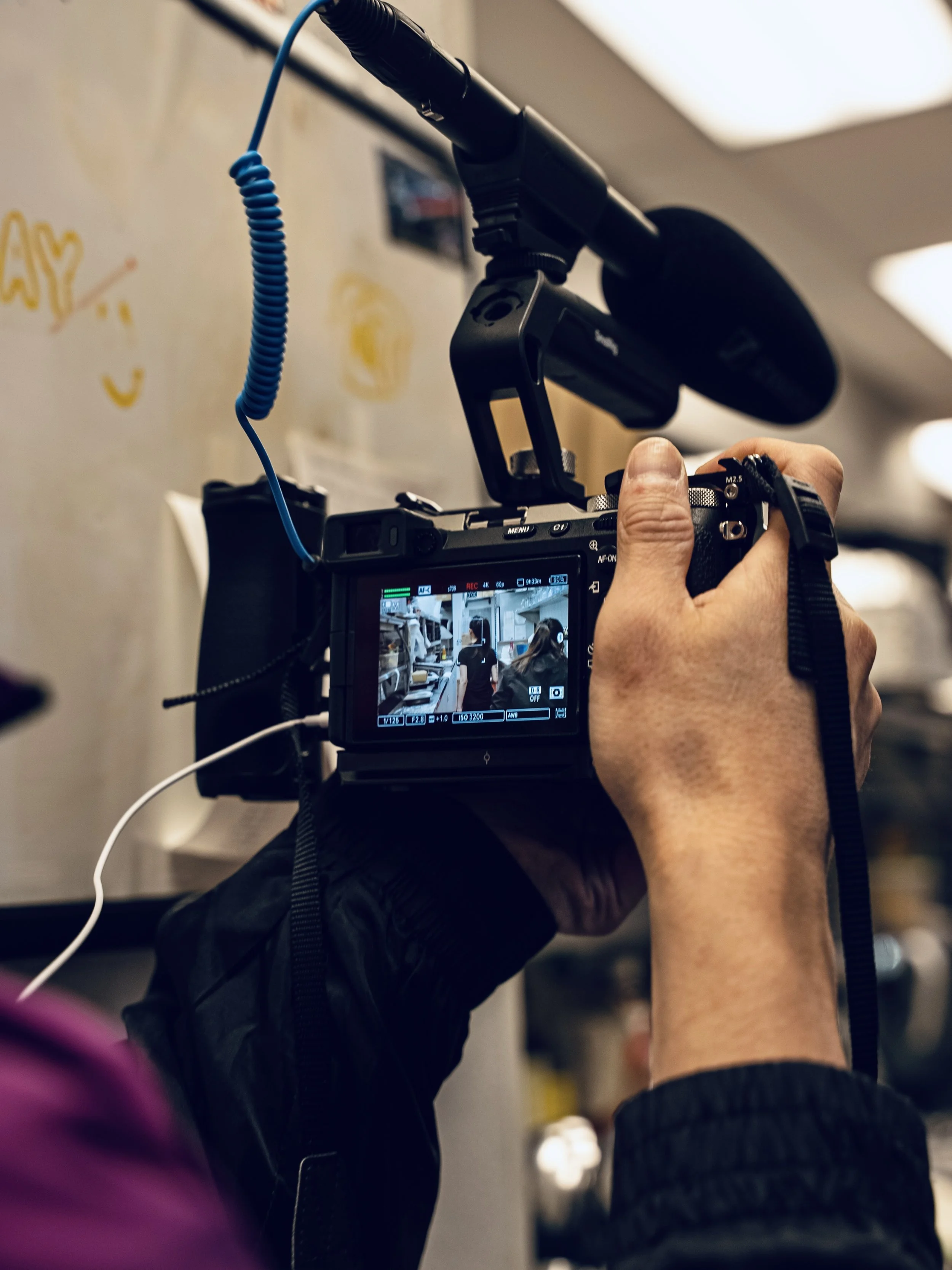 A person adjusting a camera mounted on a shoulder rig in an indoor setting, with multiple individuals visible in the background on the camera's display.