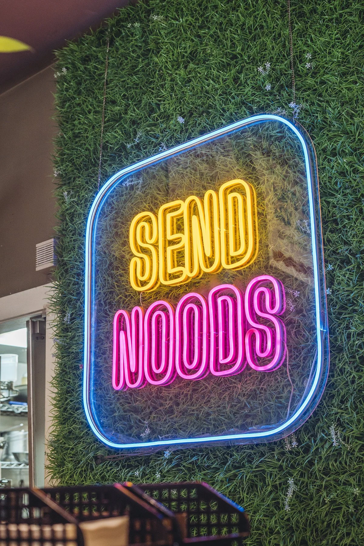 Neon sign reading 'SAND NOODLES' mounted on a leafy green wall inside a restaurant or cafe.