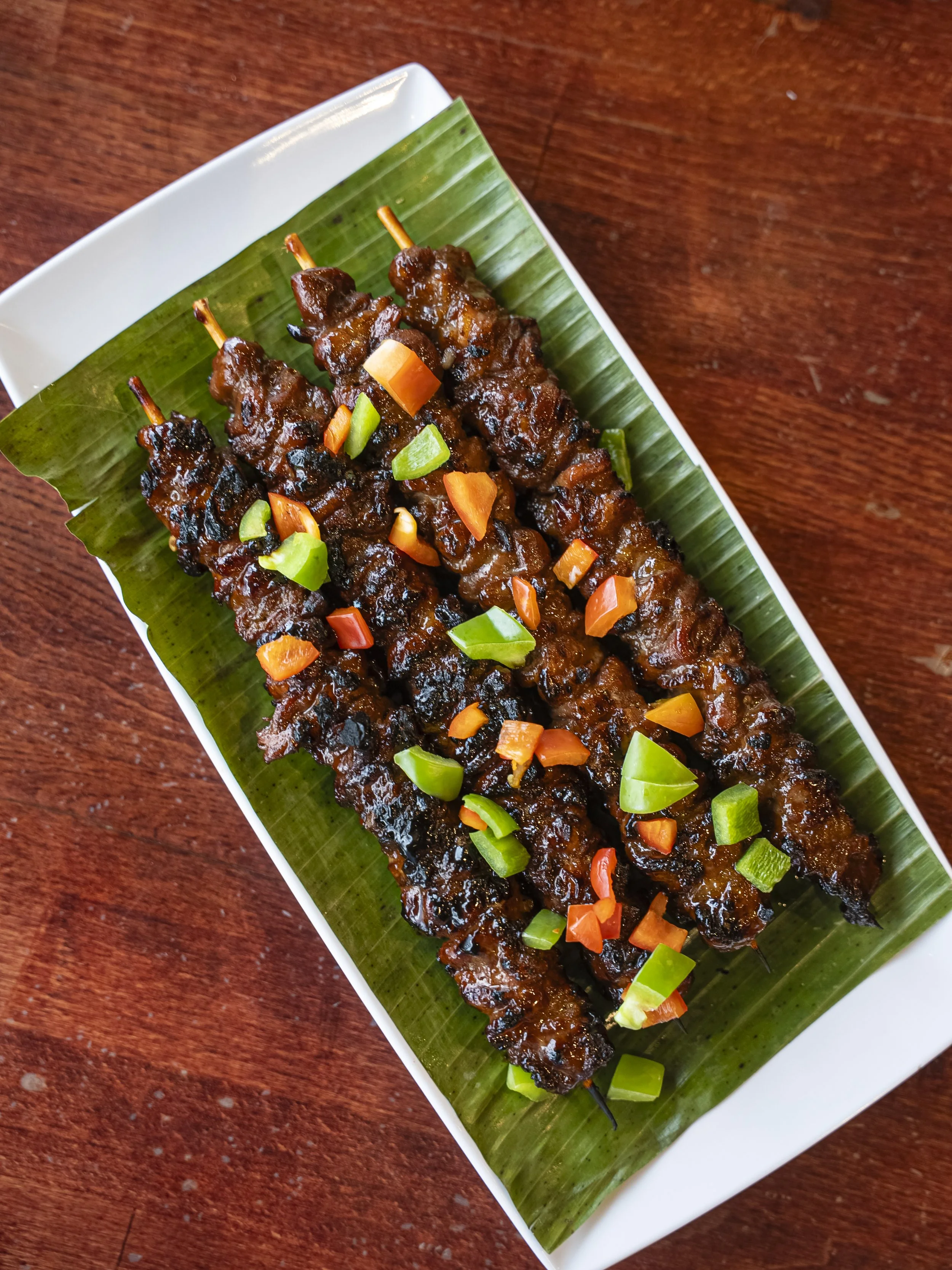 Skewers of grilled meat on a white plate with a green banana leaf, garnished with chopped green and orange peppers, on a wooden table.