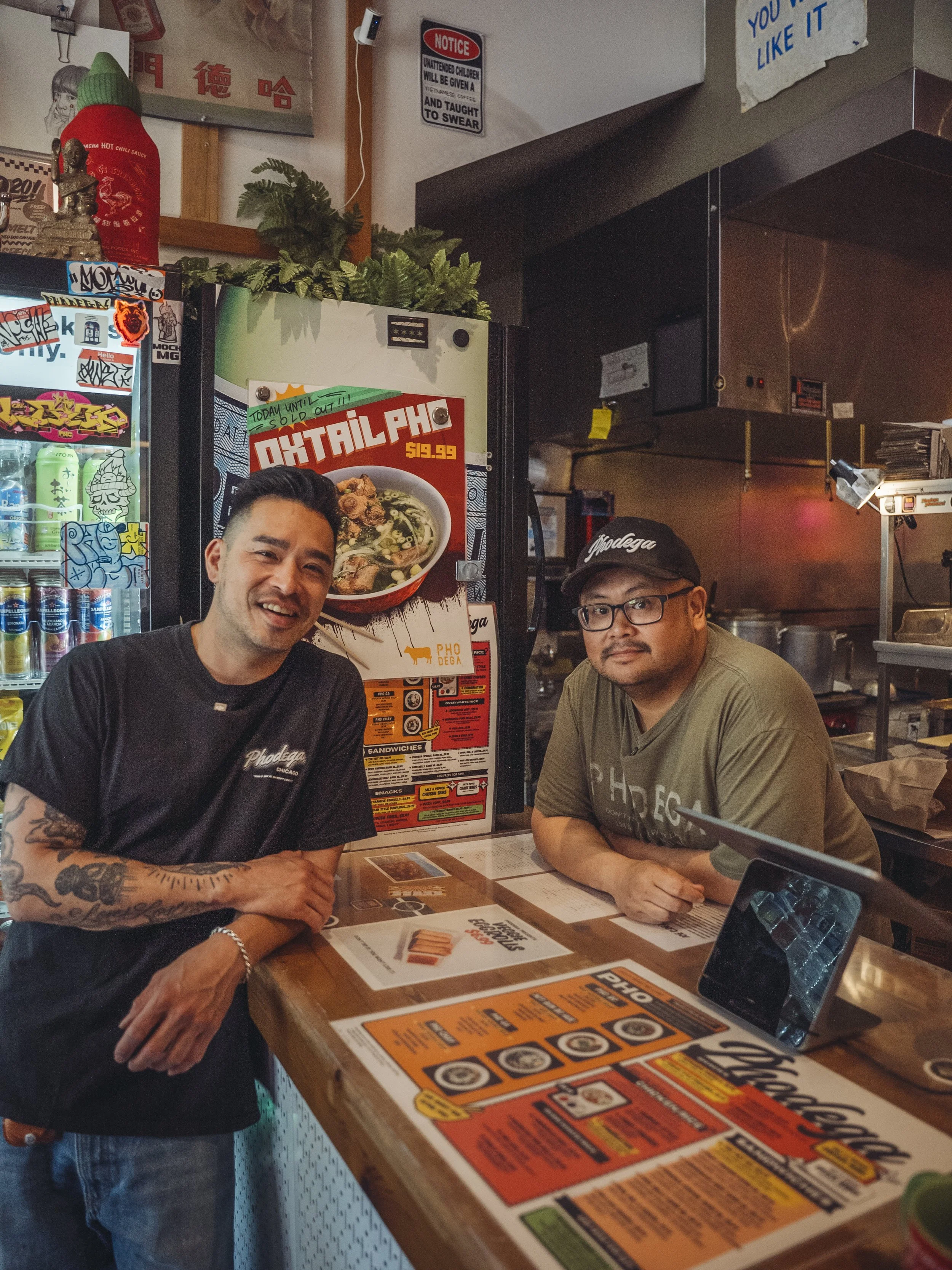 Two men stand at a restaurant counter, smiling and looking at the camera. The man on the left has tattoos on his arms and is wearing a black t-shirt. The man on the right is wearing glasses, a cap, and a gray t-shirt. The background shows a menu and various restaurant decorations.
