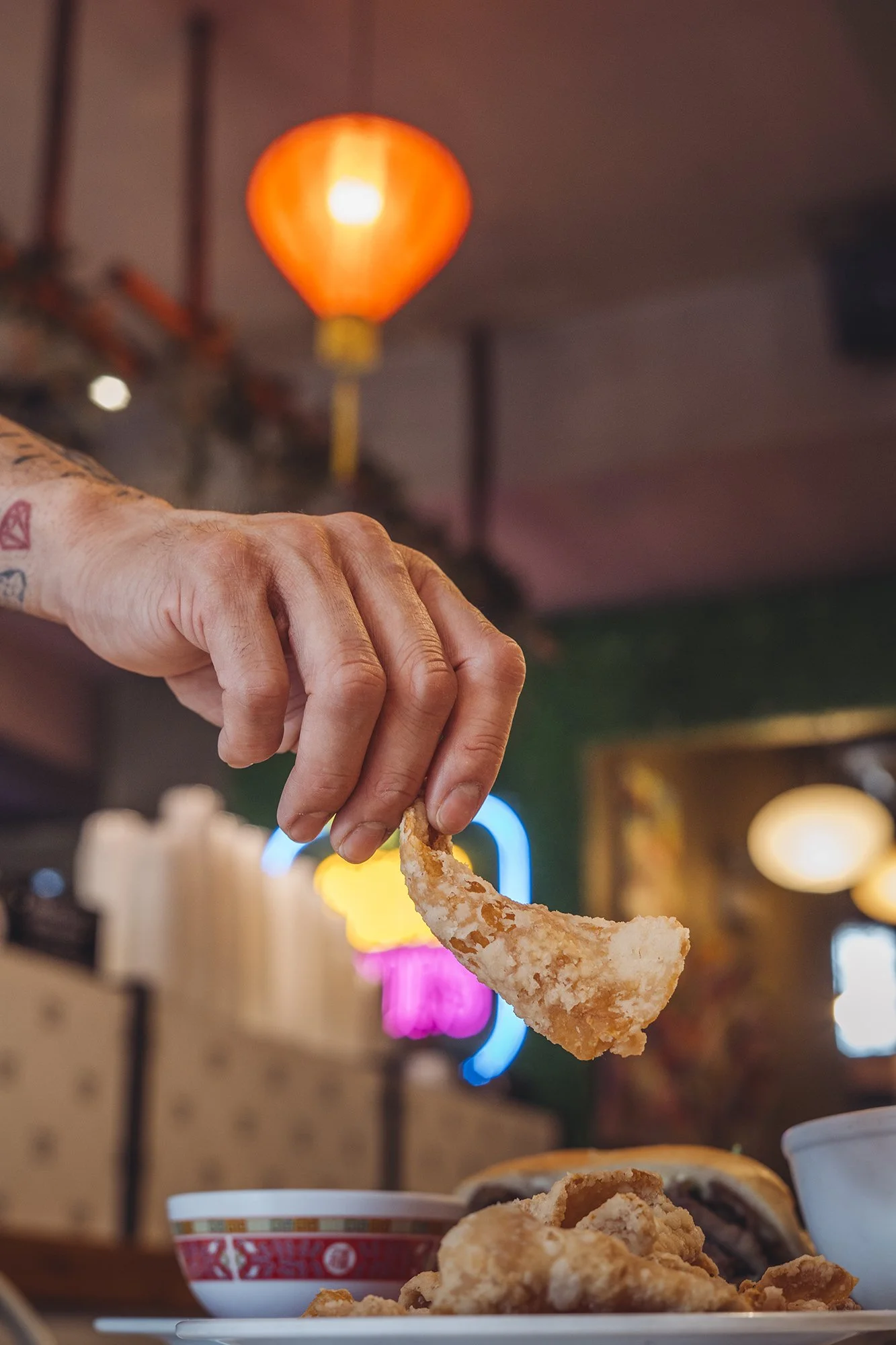 A person's hand holding a piece of fried food over a table with more fried food, a bowl of rice, and a bowl of soup in the background, in a restaurant with neon signs and hanging orange lamp lights.