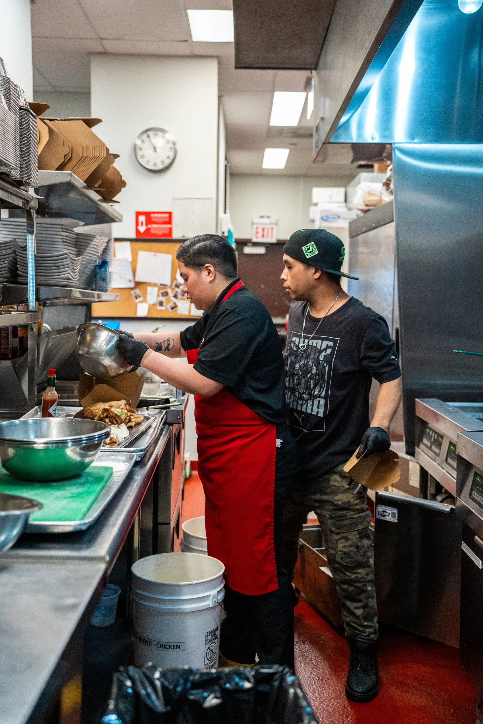 Two men working in a commercial kitchen, one wearing a red apron and black gloves, the other in a black T-shirt, camouflage pants and a backwards cap, preparing food at a stainless steel counter with various kitchen items around.