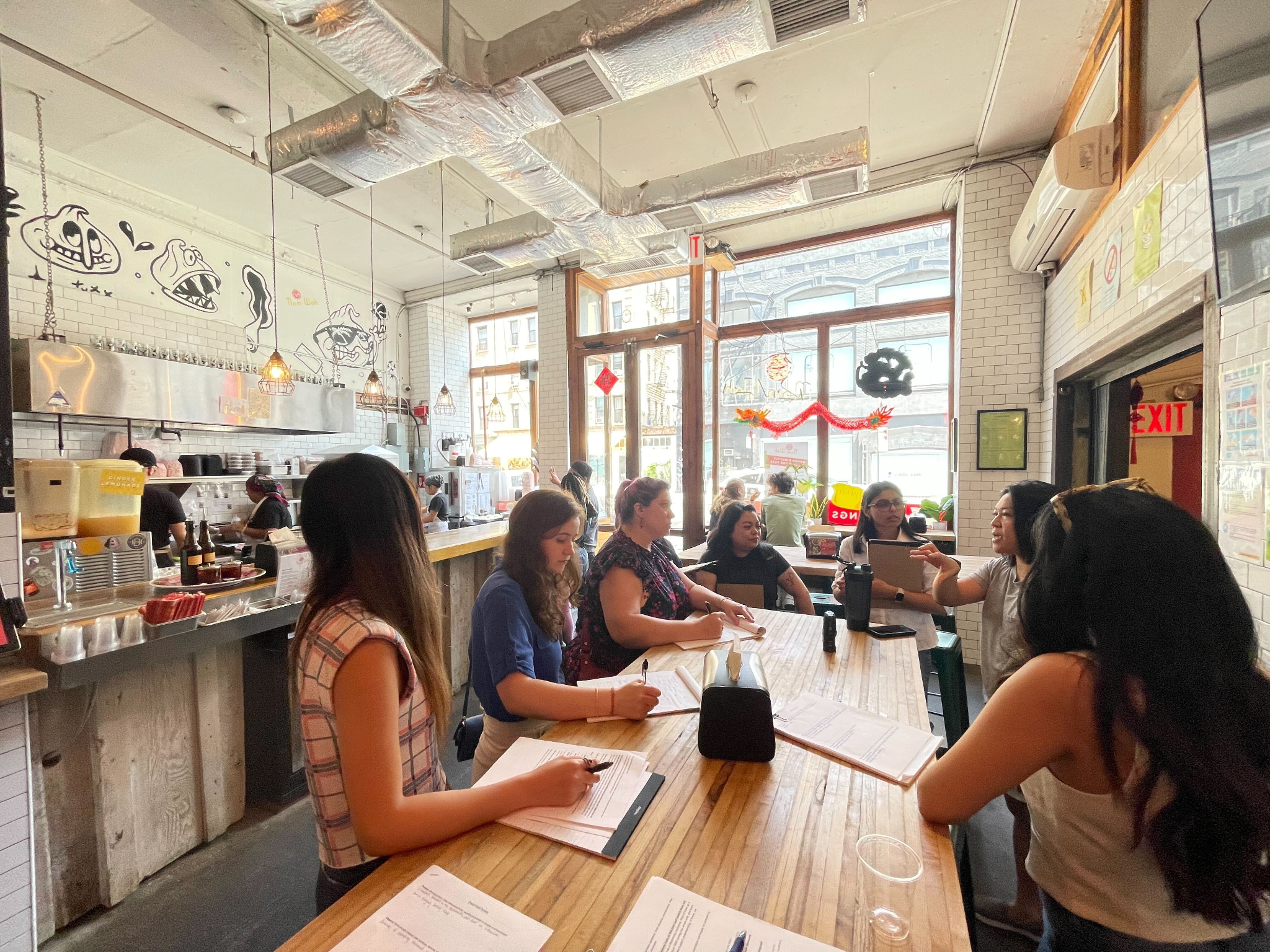 Designers sitting at a long wooden table inside a lively restaurant with large windows, white brick walls, hanging lights, and decorative artwork, engaging in conversation with an employee.