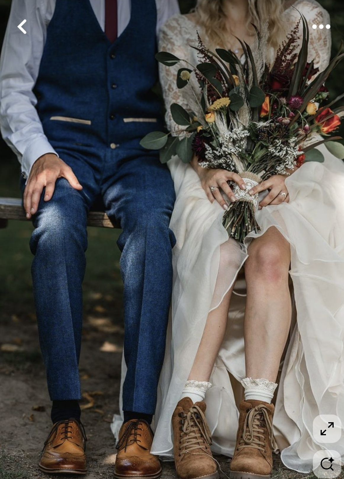 Dettaglio di due persone sedute su una panchina, una in abito da sposa e l'altra in abiti casual, con scarpe marroni e una mano che tiene un mazzo di fiori.