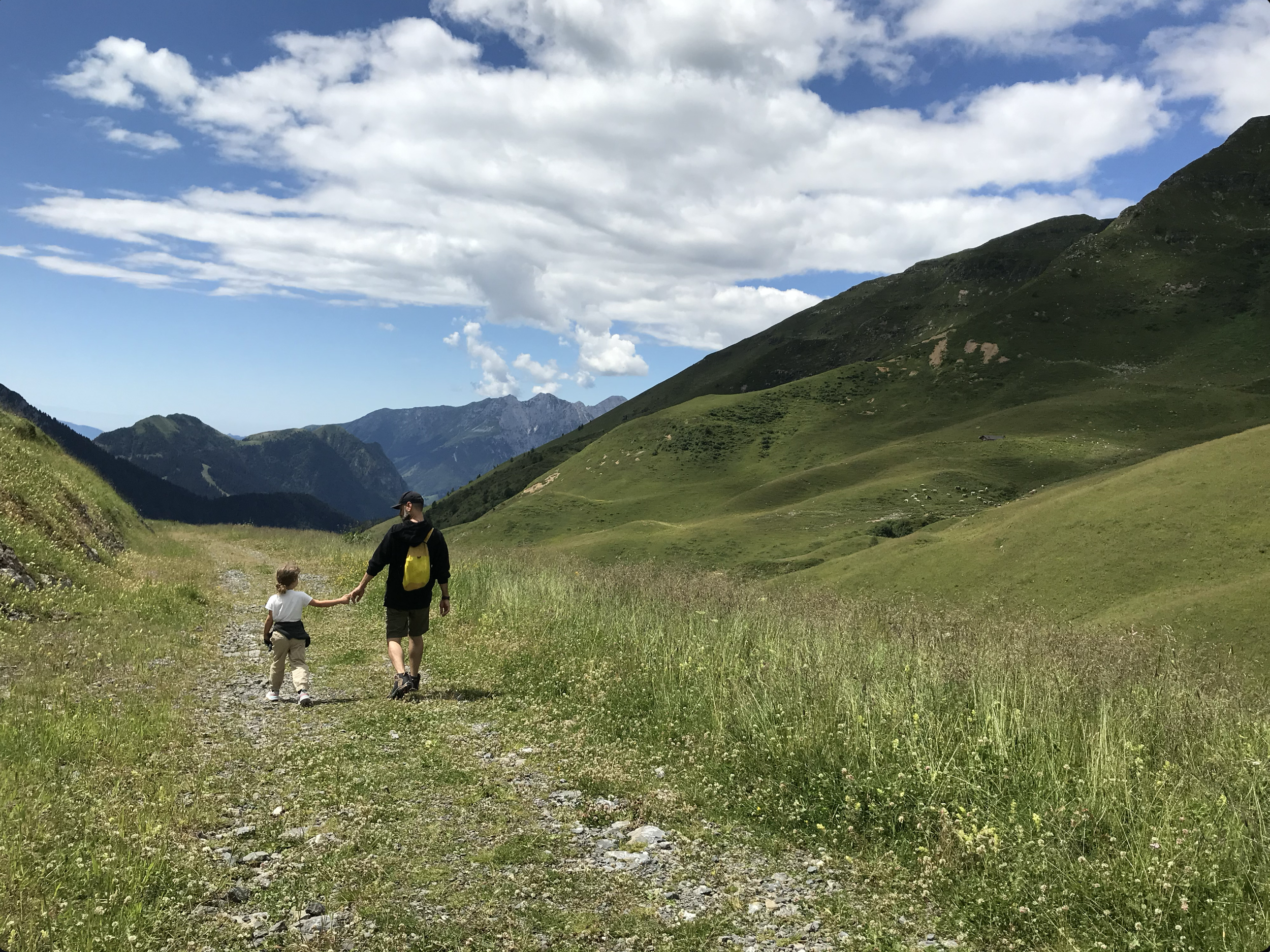 Due persone, un adulto e un bambino, camminano lungo un sentiero in una valle montuosa verde sotto un cielo con nuvole.