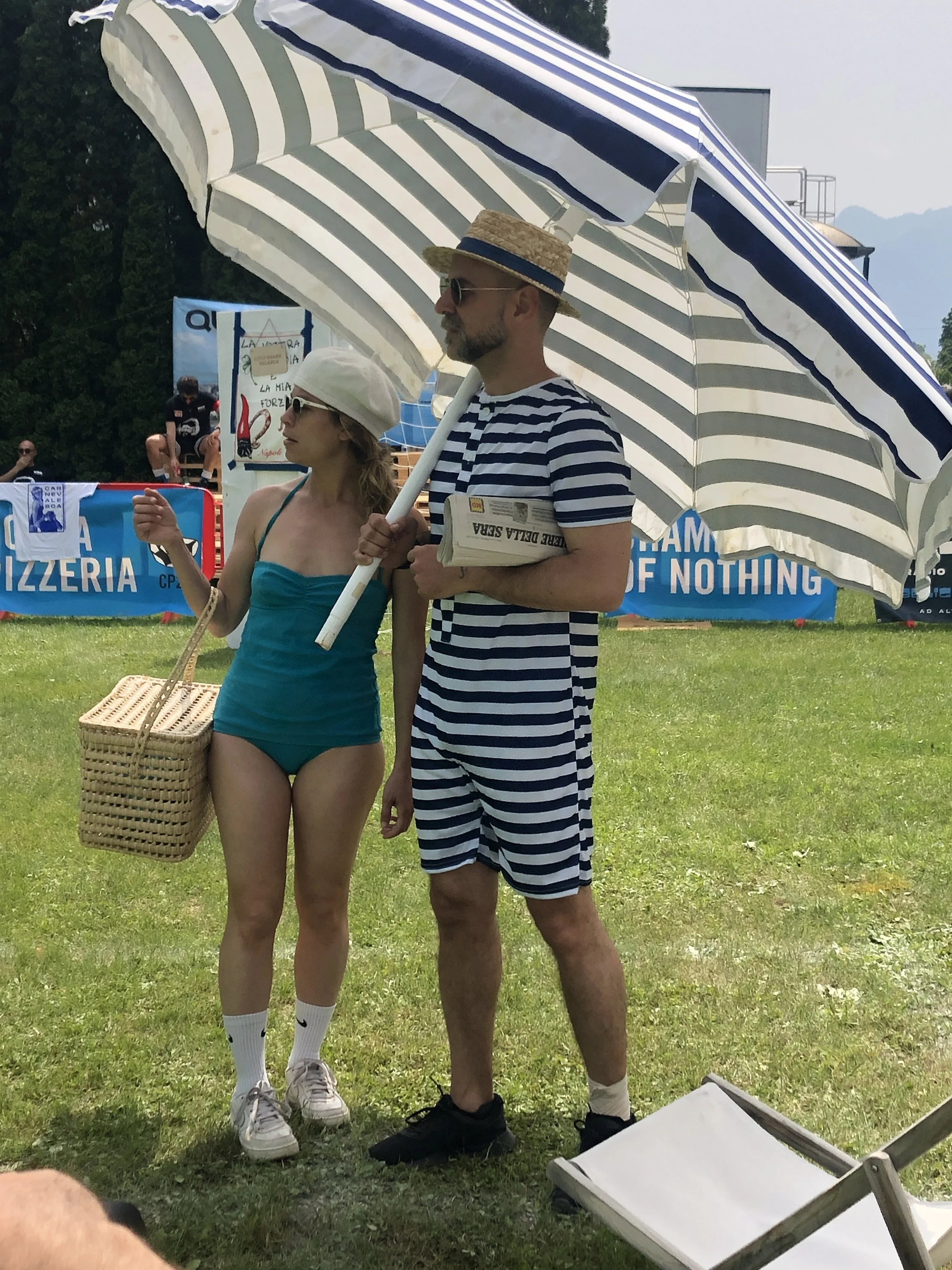 Due persone in spiaggia sotto un grande ombrellone, un uomo con vestiti a righe e un cappello di paglia, una donna con vestiti da bagno, entrambi con occhiali da sole, parlano mentre tengono oggetti in mano. Sullo sfondo ci sono altre persone, banchine e cartelloni pubblicitari.