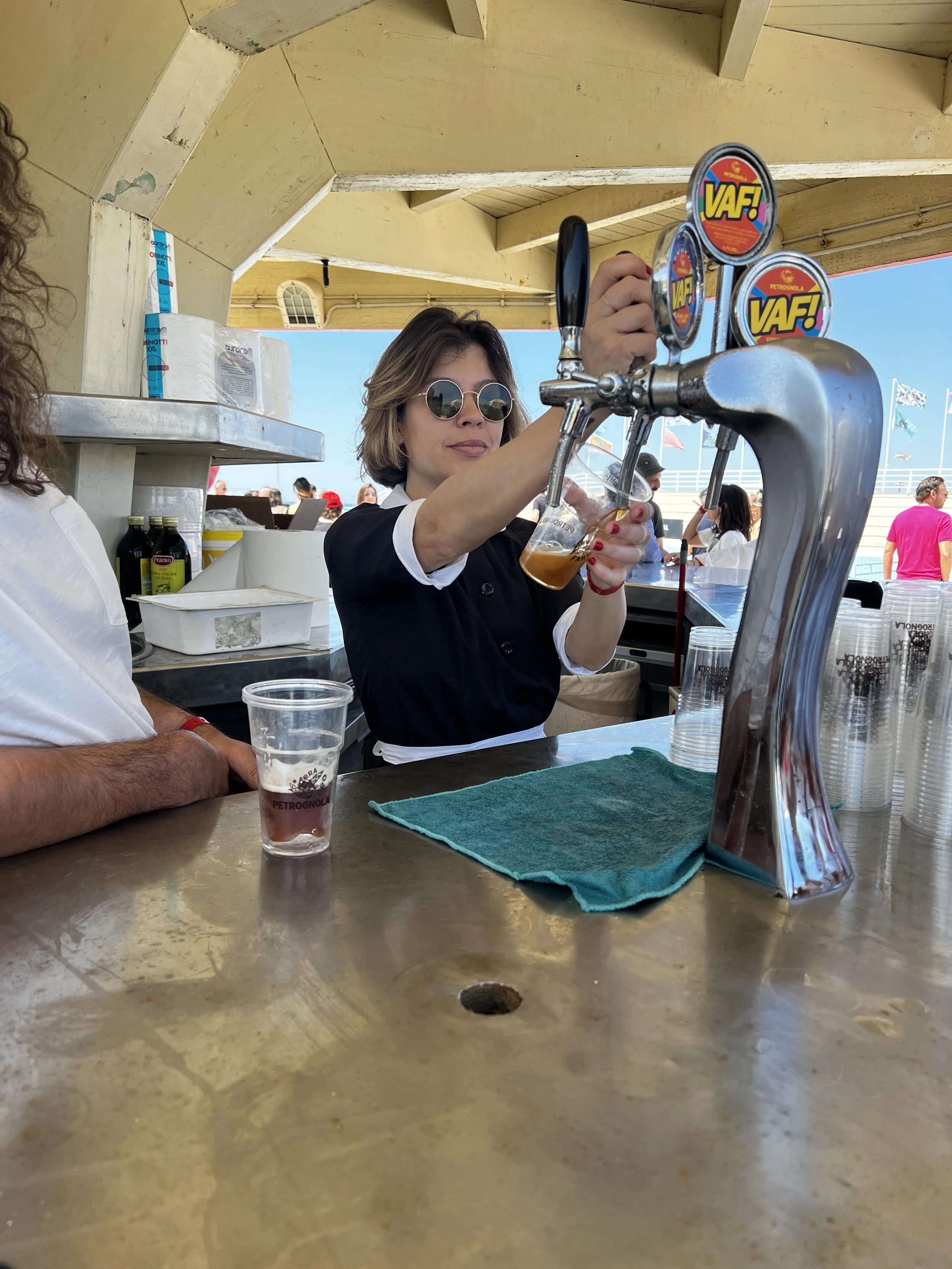 Una donna con occhiali tondi versa birra da un rubinetto in un chiosco da bar all'aperto.