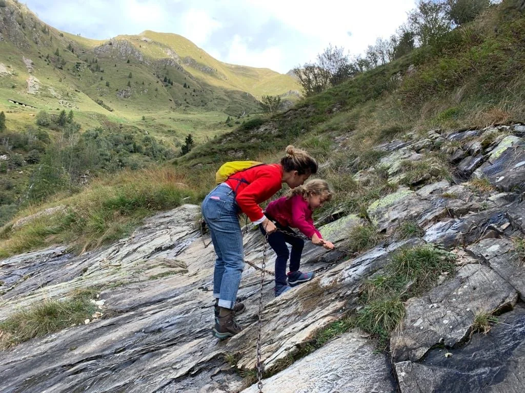Una donna e una bambina stanno arrampicandosi su una roccia vicino a un ruscello in un paesaggio montano verde.