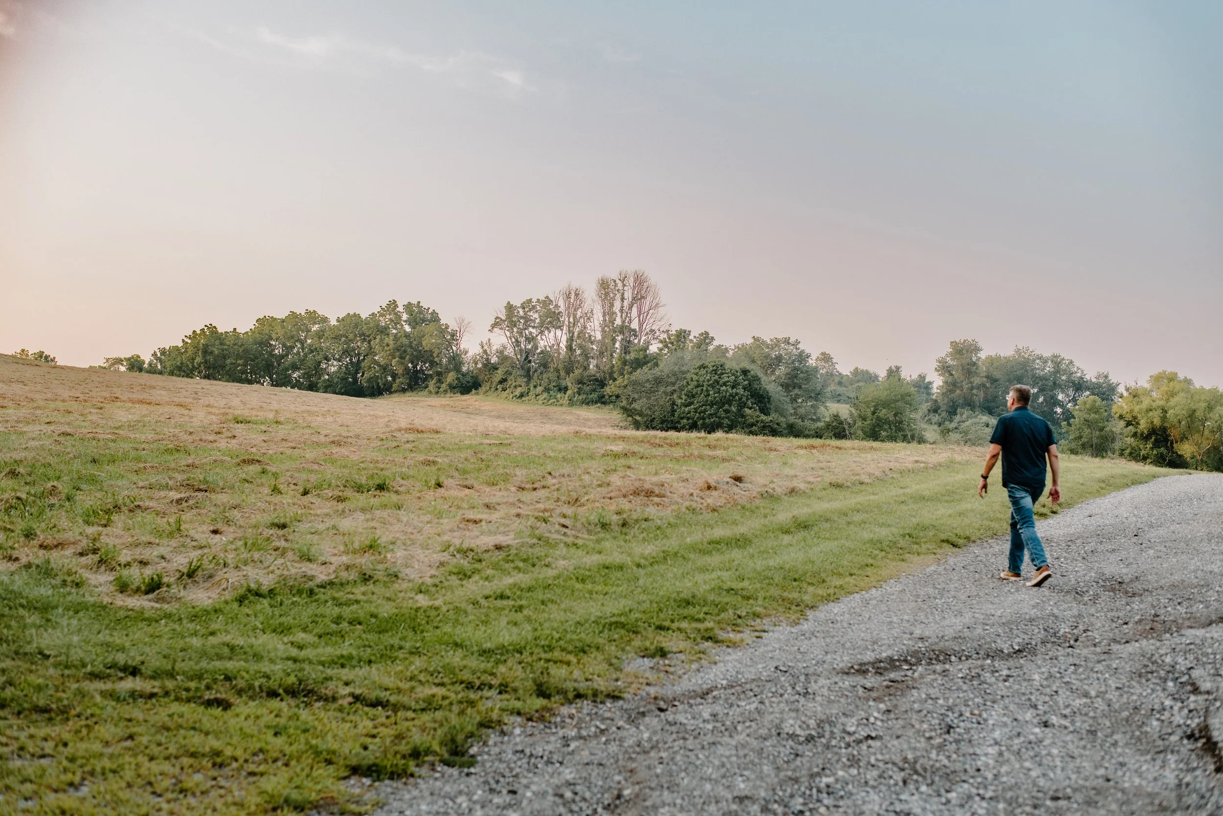 Marc Lucenius walking on a gravel trail in a rural grassy field with trees in the distance, under a partly cloudy sky.