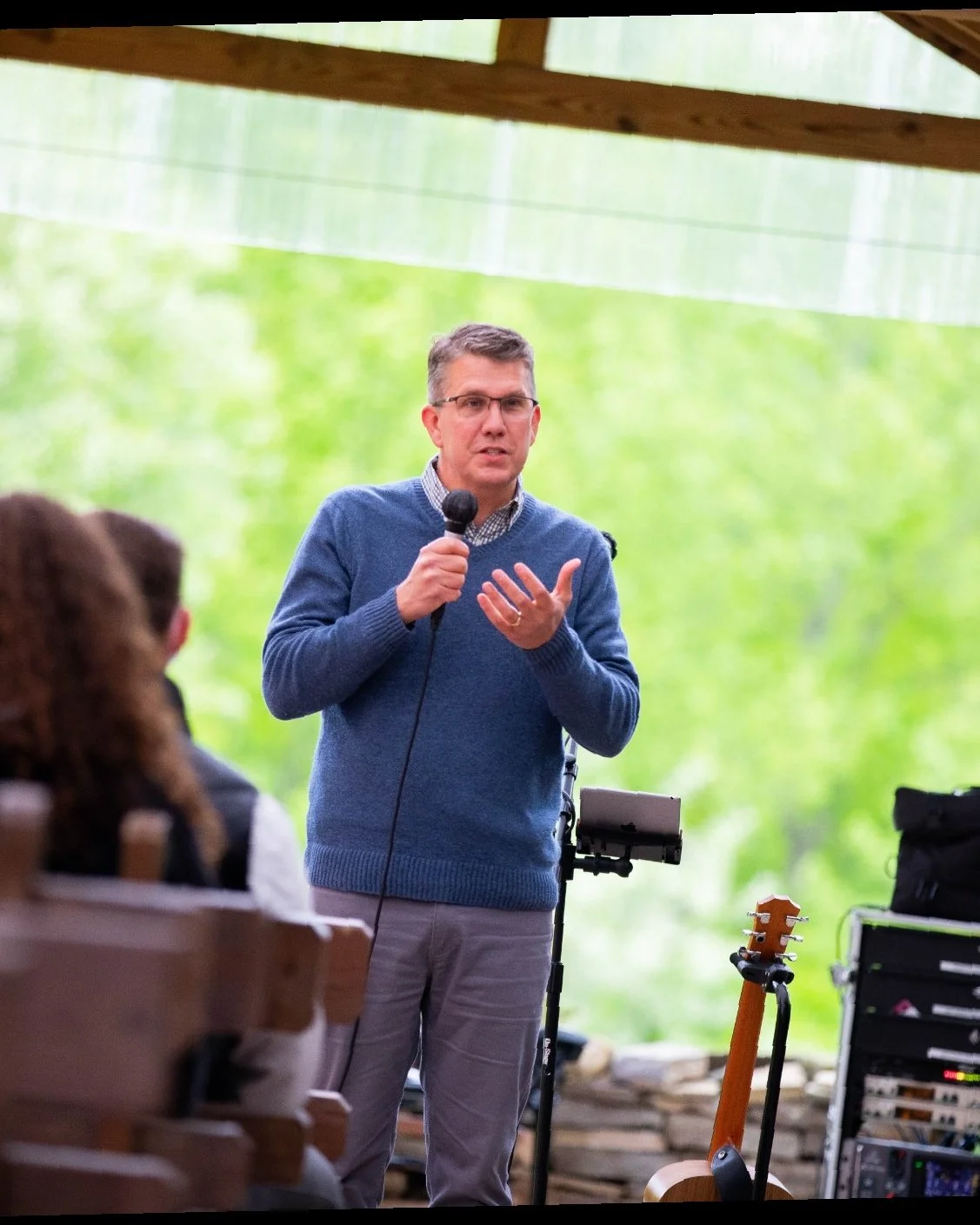 Marc Lucenius in a blue sweater and glasses speaking into a microphone during an outdoor event, with a blurred audience in the foreground and green trees in the background.