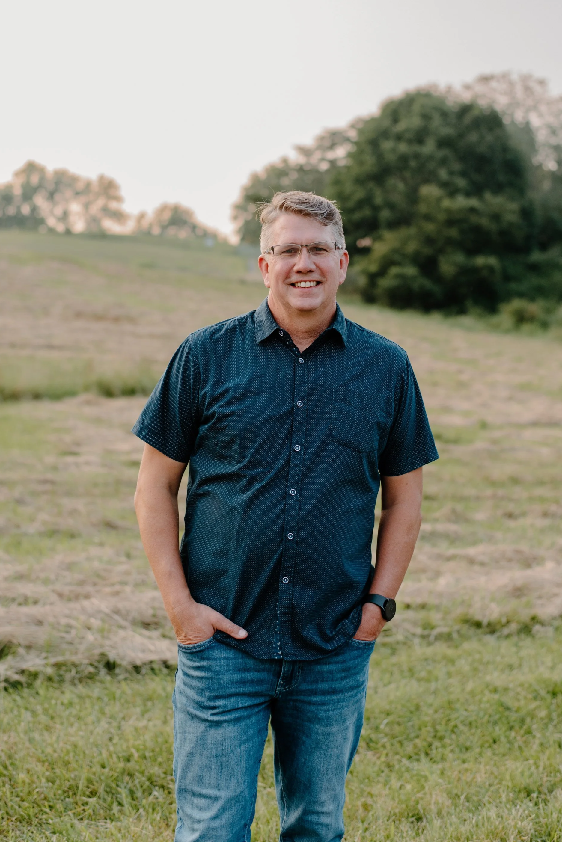 Smiling man wearing glasses, dark blue shirt, and jeans standing outdoors in a grassy field with trees in the background.