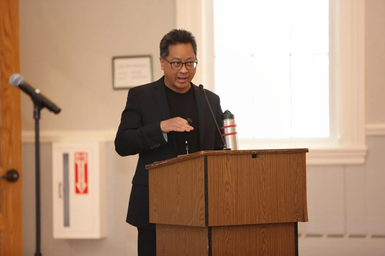 A man wearing glasses and a black suit speaking at a wooden podium with a microphone, in a well-lit room with a window and framed certificate on the wall.