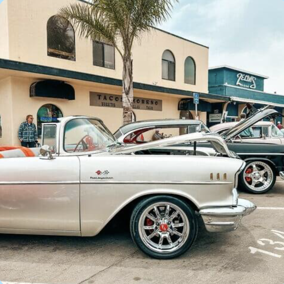 A line of vintage cars parked on the street in front of a restaurant with people nearby. A palm tree is in the foreground.