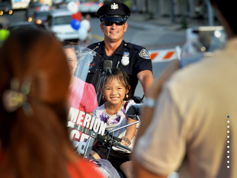 Young girl smiling while sitting on a police motorcycle, with a police officer and others around during a community event in daylight.