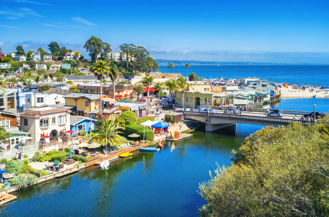 A coastal town with colorful houses, palm trees, and a bridge over water, leading to a beach with umbrellas and people, ocean in the background on a sunny day.