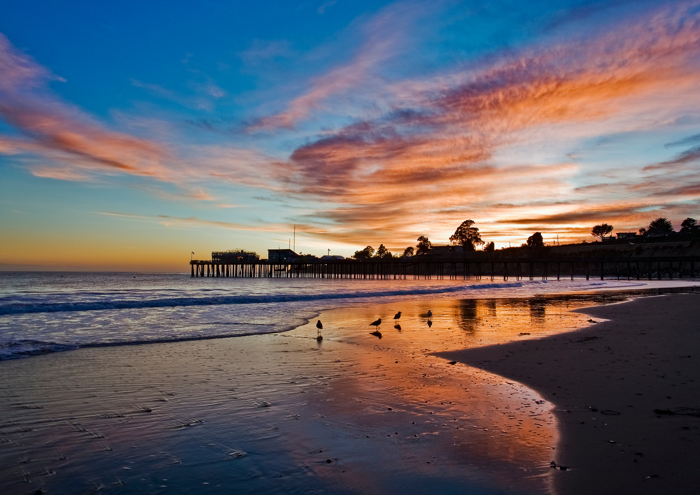 Sunset over a beach with a pier extending into the ocean, reflecting colorful sky and clouds, with seagulls along the shoreline.
