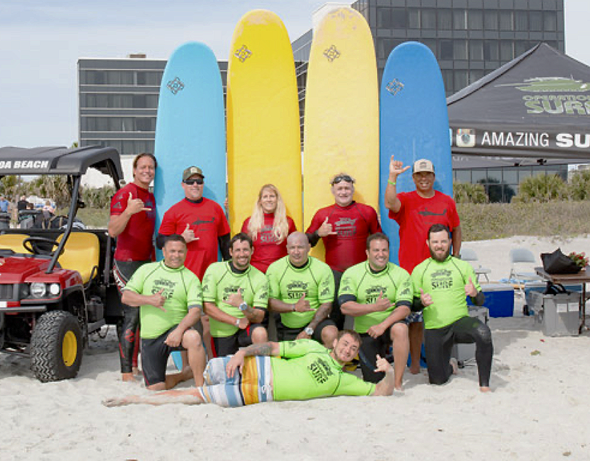 Group of people at the beach with colorful surfboards, a golf cart, and a tent in the background, some making a thumbs-up gesture.